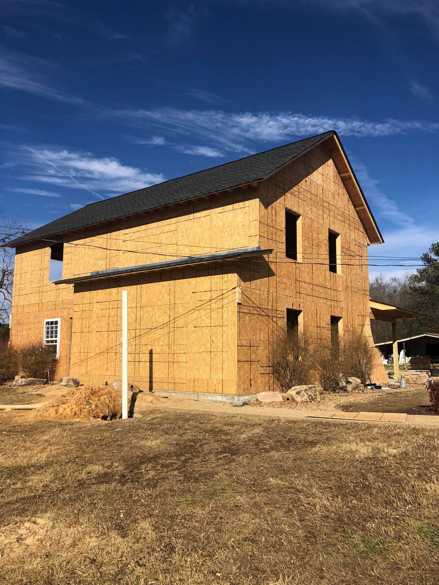 Two-story house under construction with exposed wooden frame. Blue sky, unfinished windows and dark roof.