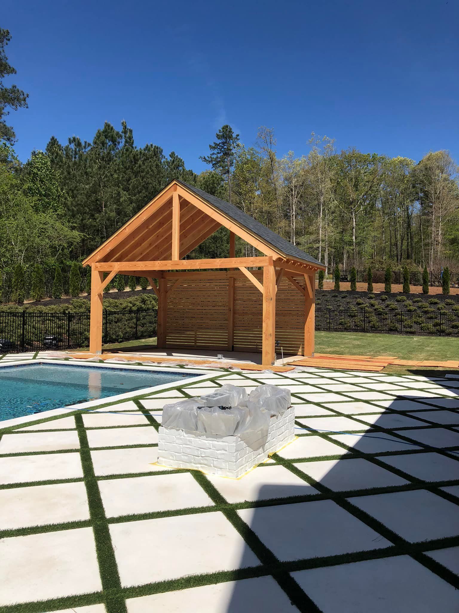 Wooden pool cabana with blue pool, concrete patio, and trees under a clear sky.