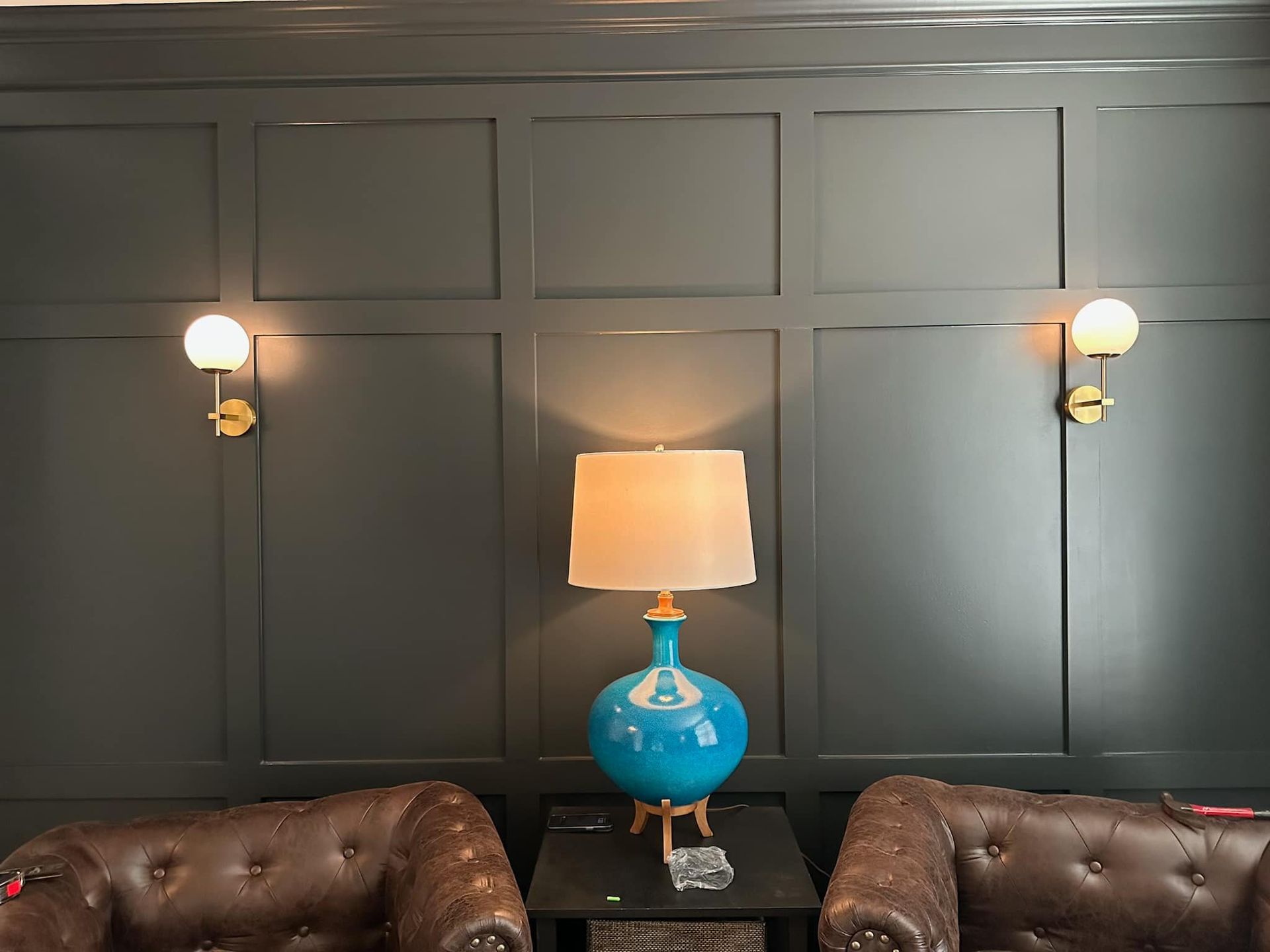 Dark green paneled wall with a teal lamp, two sconces, and two brown leather armchairs.
