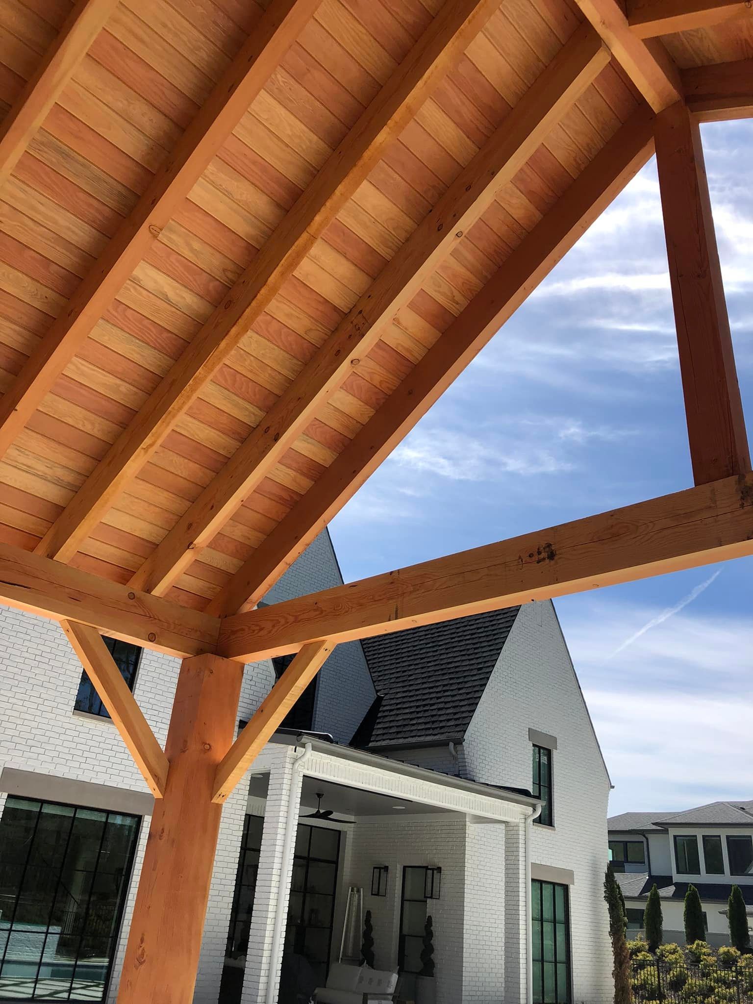 Wooden patio roof and beams with a view of a white brick house and sky.