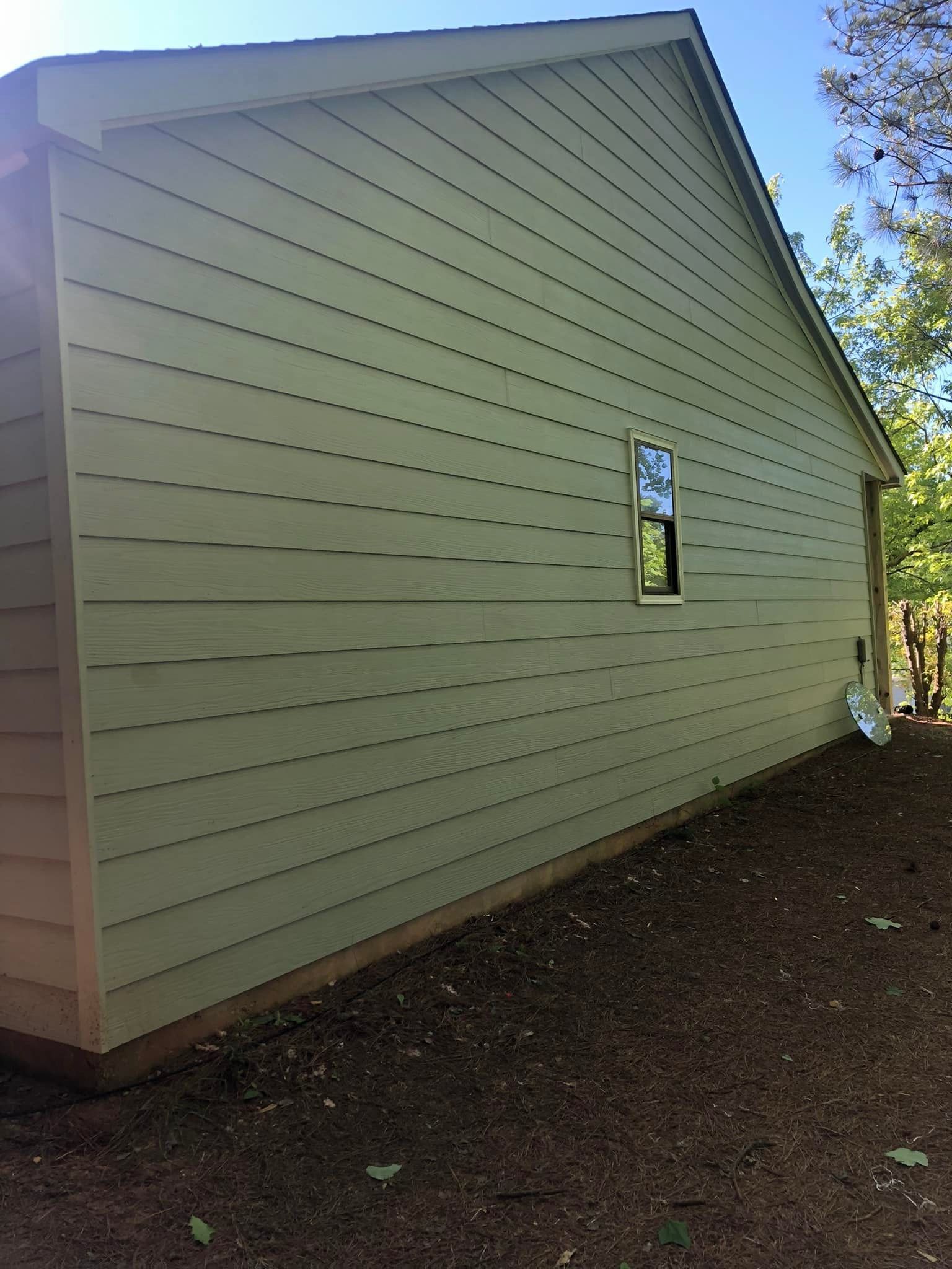 Green-sided building with small window and angled roof, set in a yard with dirt and trees.
