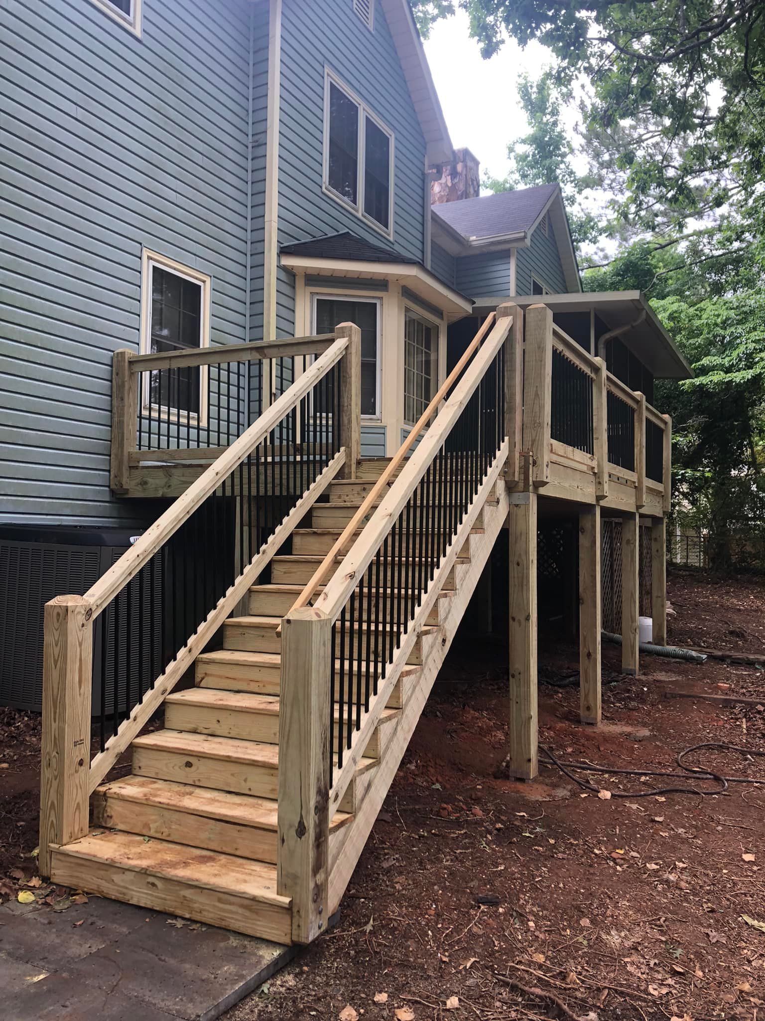 Wooden deck with stairs leading to a light blue house. Black metal railings and surrounding trees are visible.
