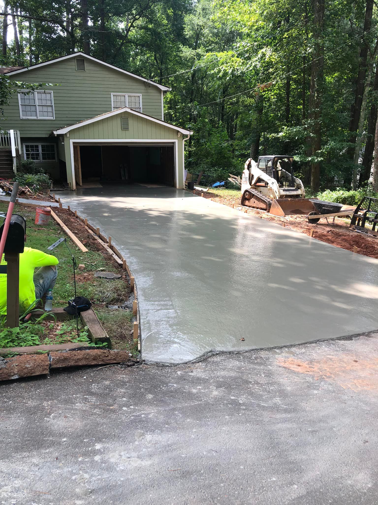 Freshly poured concrete driveway in front of a green two-story house with a garage.  An excavator is in the background.