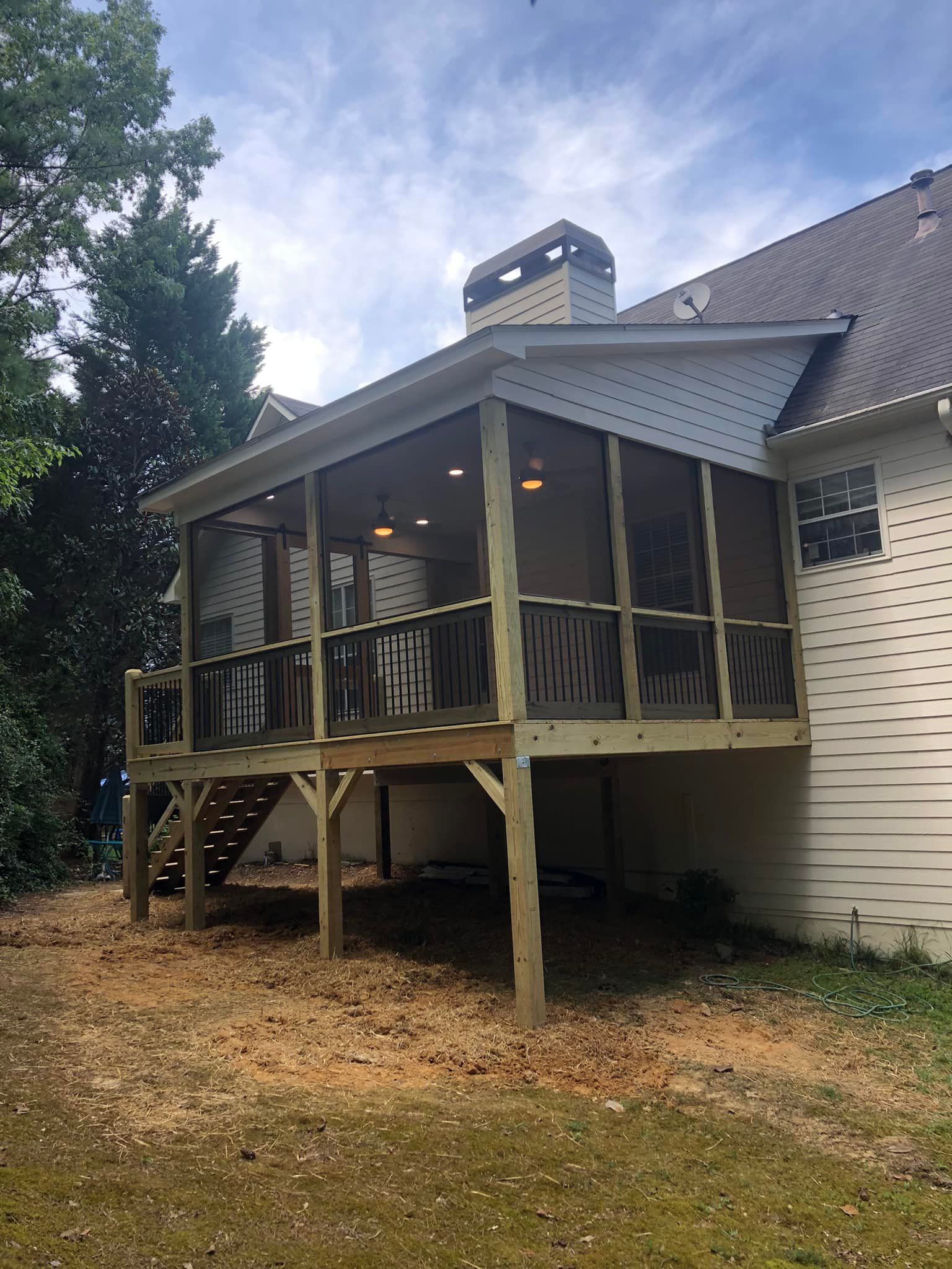 Screened-in porch addition with wood deck, dark wood railing, and roof against a white house and blue sky.