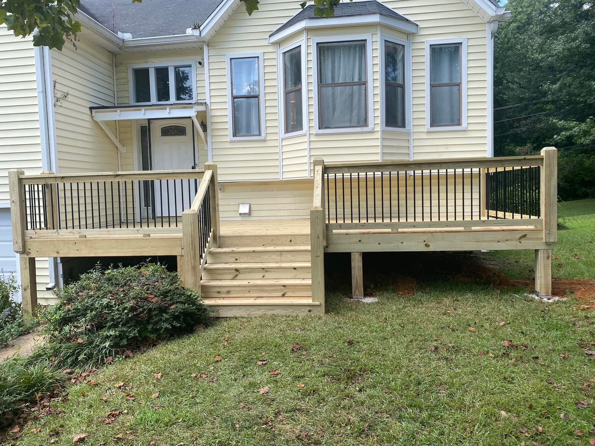 A newly constructed wooden deck with stairs in front of a light yellow house with a bay window.