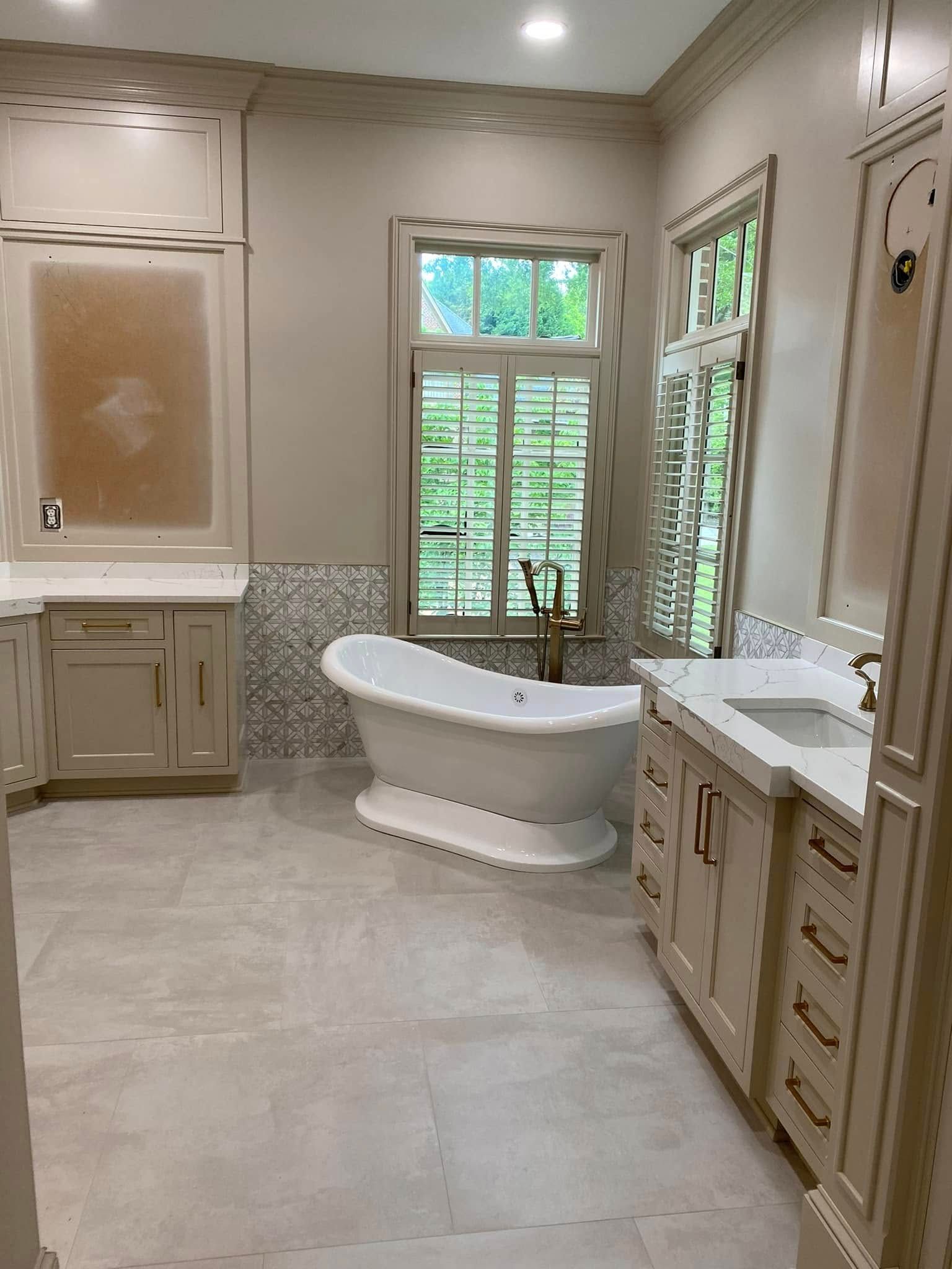 Elegant bathroom with a free-standing tub, two vanities, a window with blinds, and light-colored cabinetry.