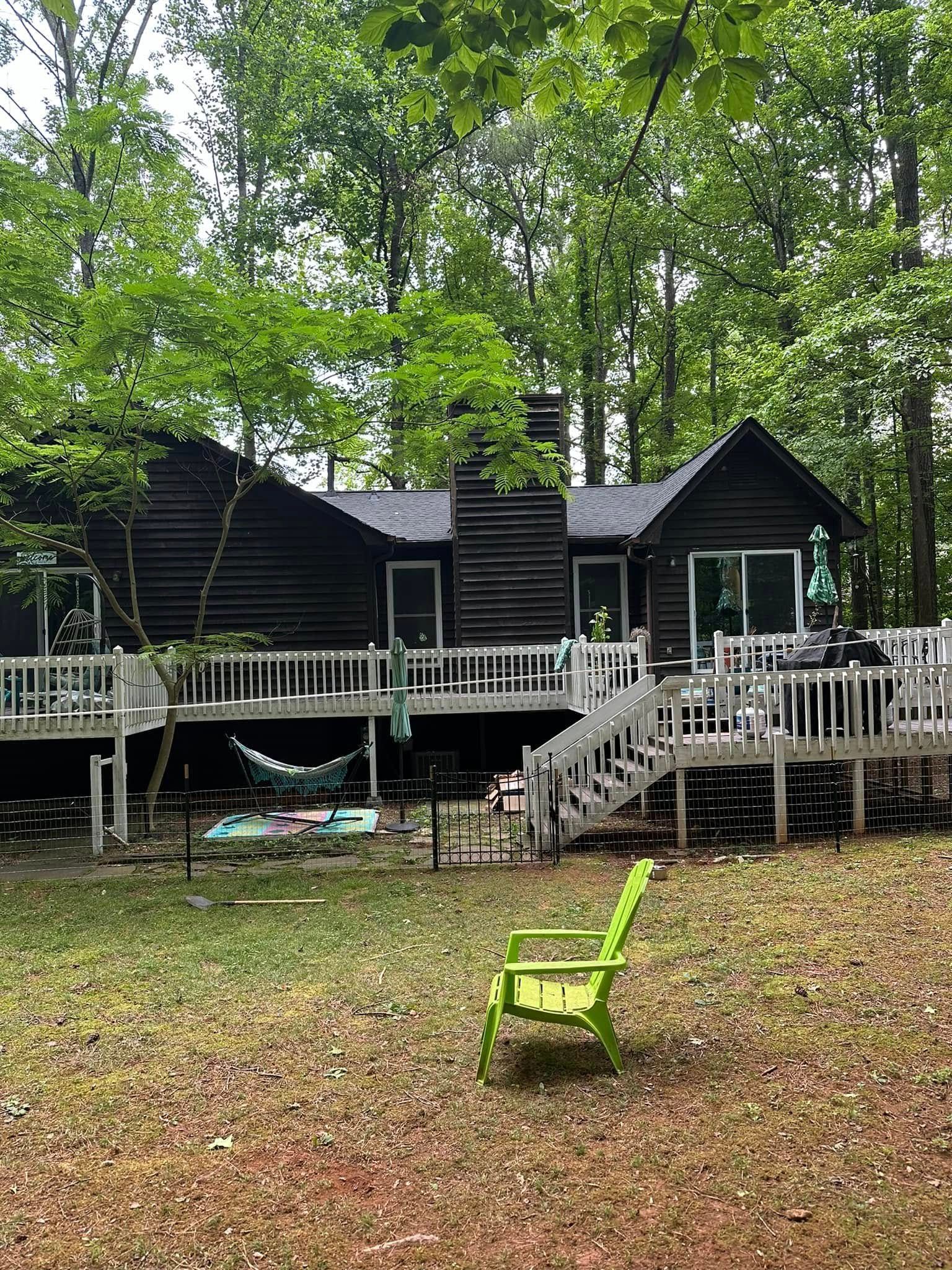 Dark cabin with white deck in a wooded area, green chair in the yard.