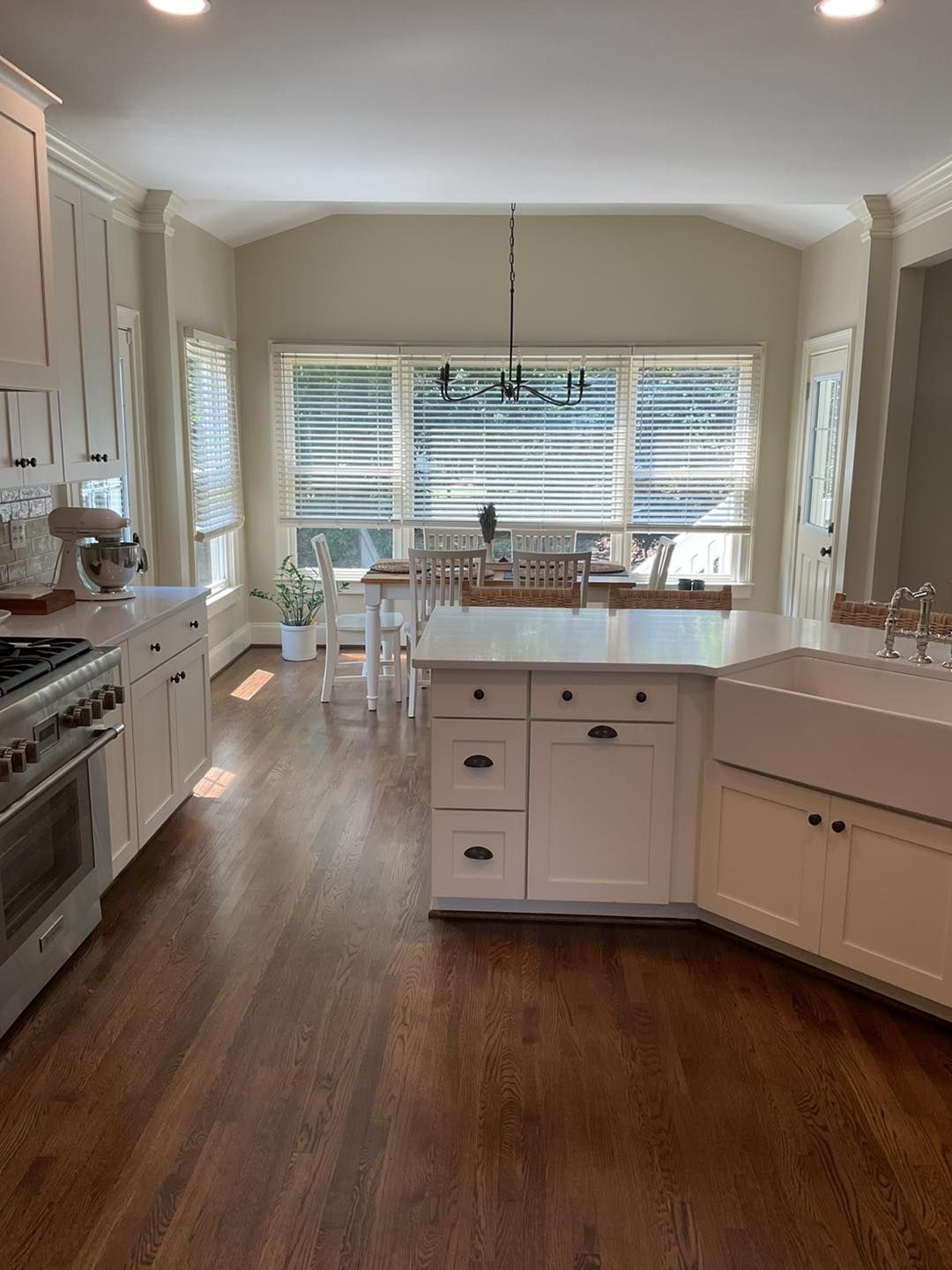Bright kitchen with white cabinets, island, and appliances. Wooden floor and large windows with blinds.