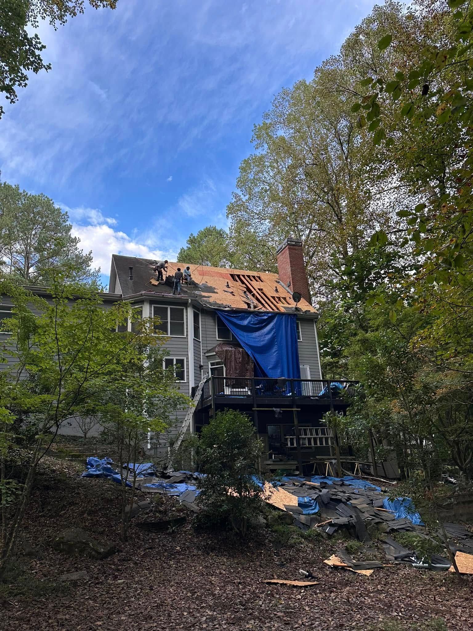 House with missing roof, covered by blue tarp; surrounded by trees, cloudy sky.