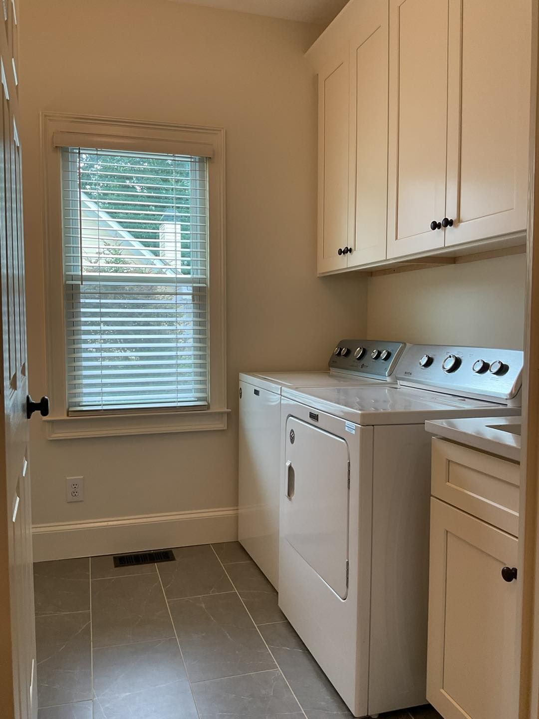 Laundry room with white washer/dryer, cabinets, and window with blinds; gray tiled floor.