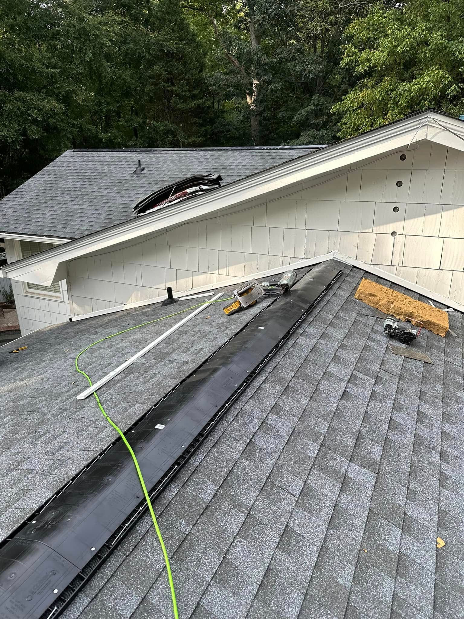 Roof with a black channel and tools, angled toward a white wall, and shaded by trees.