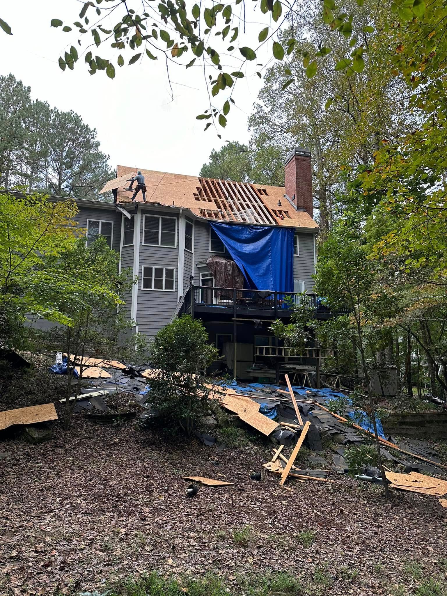 House with missing roof shingles, covered by blue tarp; surrounded by trees and debris.