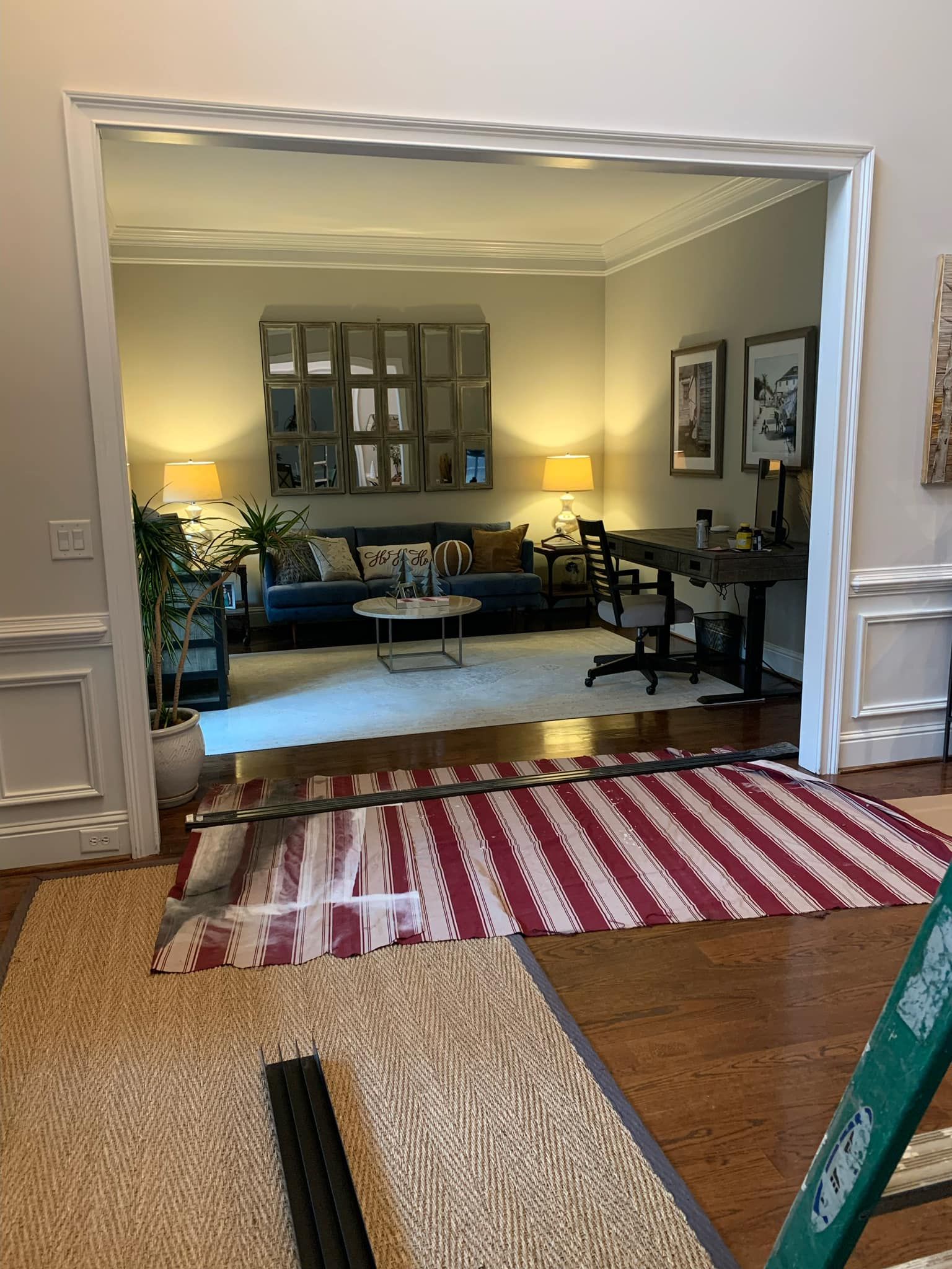 View into a living room and study area with rug, sofa, table, lamps, artwork, and desk visible.