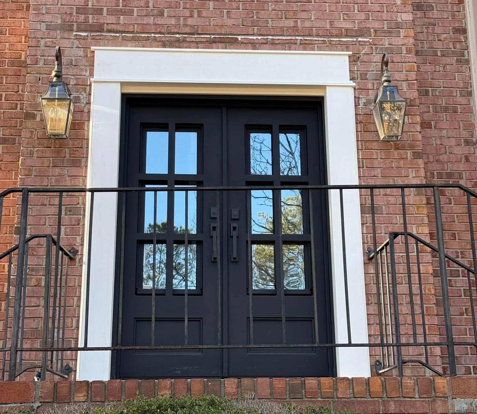 Black double doors with glass panes, flanked by lanterns, set in a brick wall with white trim and a wrought iron railing.