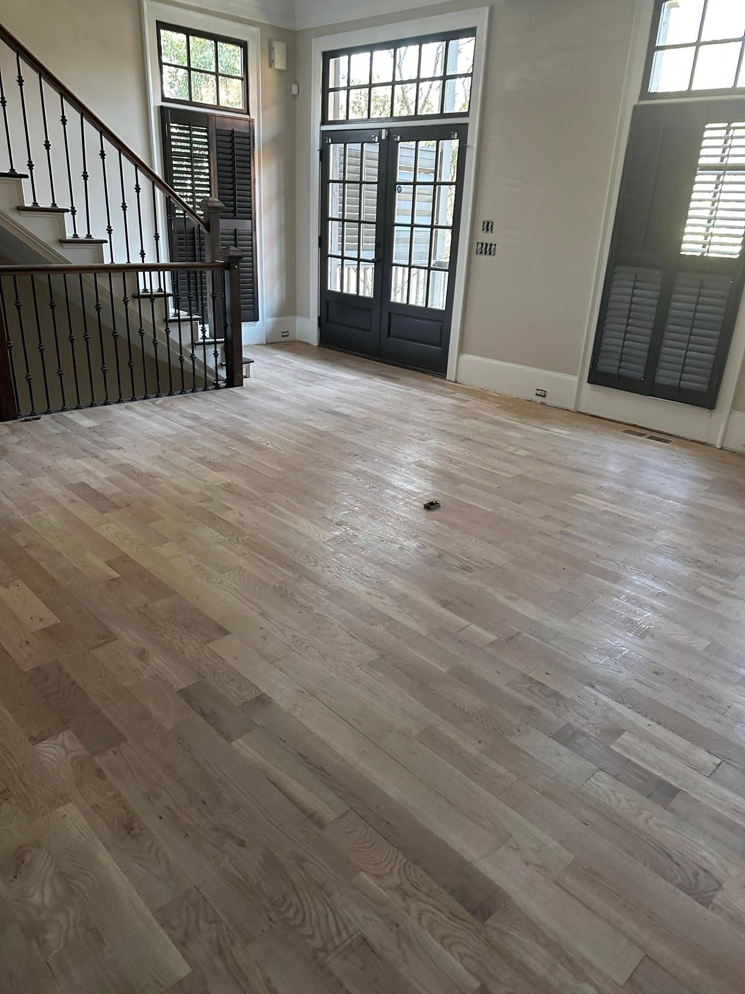 Empty room with light wood floors, stairs to the left, and a door at the back. Shutters on windows.