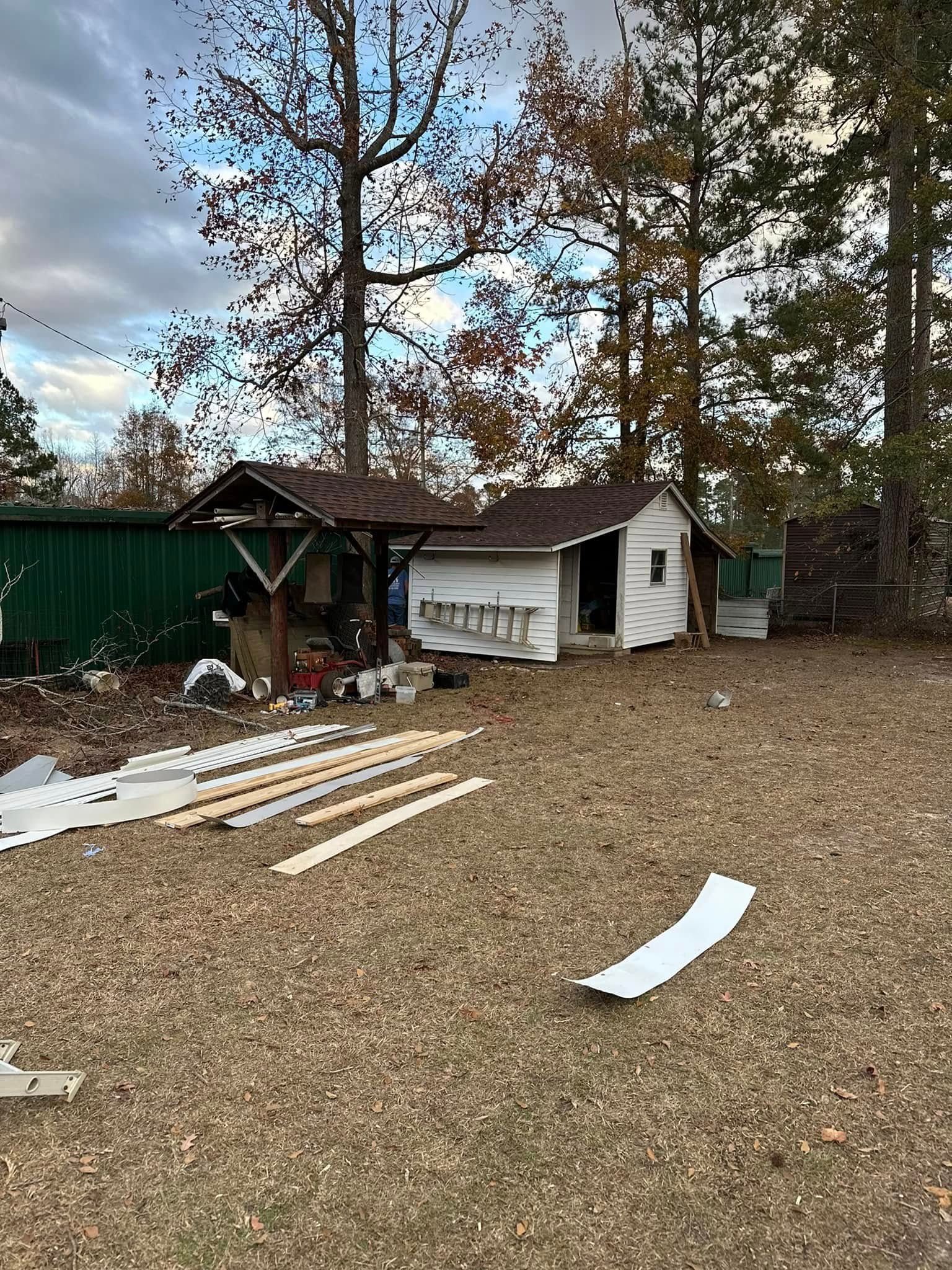A small white shed with a ladder against it, set in a yard with trees and building materials scattered about.