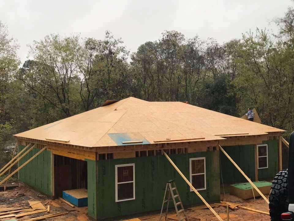 House under construction; green siding, plywood roof, supports and windows are visible, trees in the background.