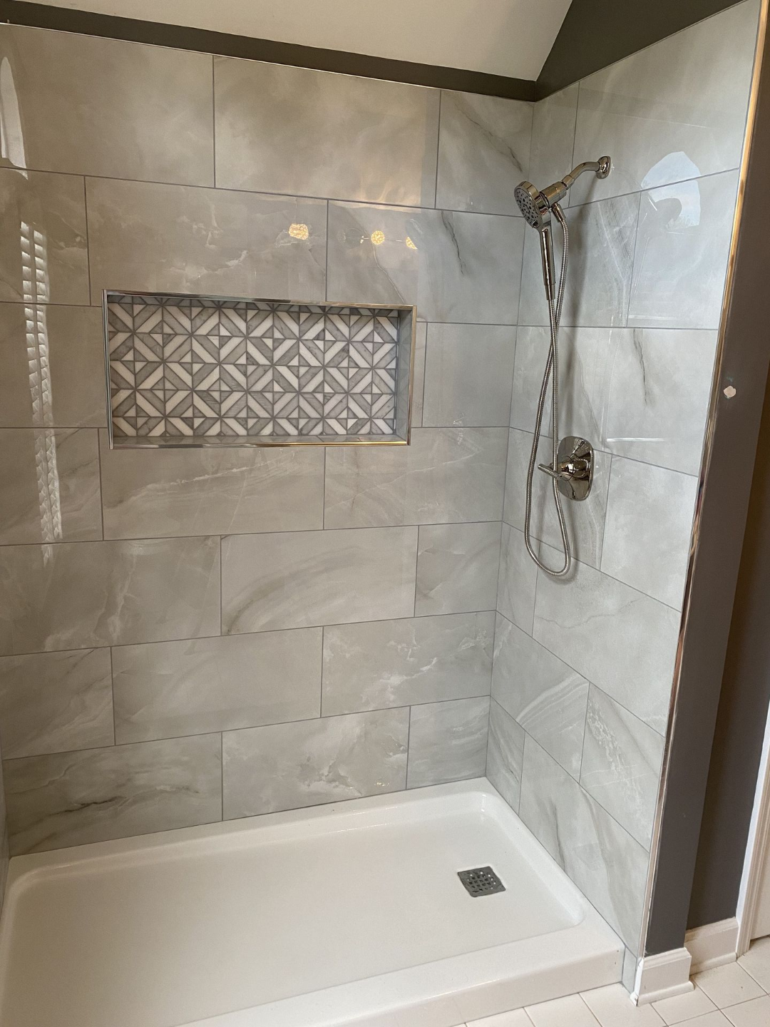 Gray tiled shower with decorative recessed shelf, chrome showerhead, and white base.