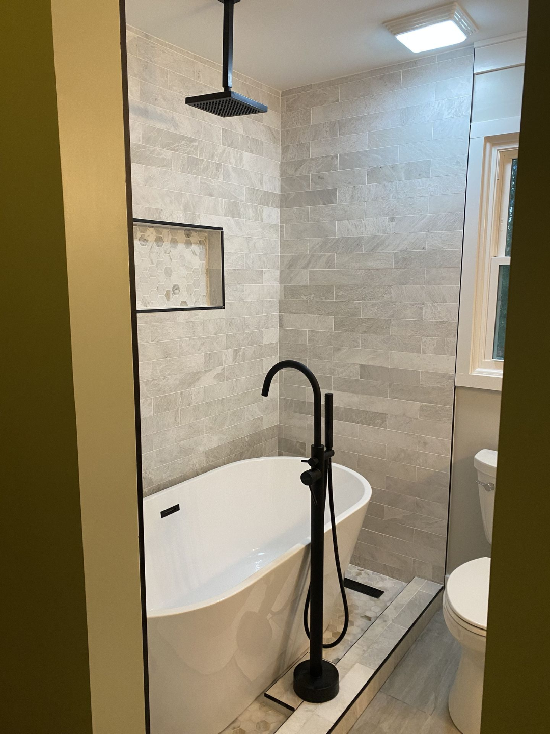 Bathroom with white tub, black showerhead/faucet, gray tiled walls, recessed shelf, and toilet.