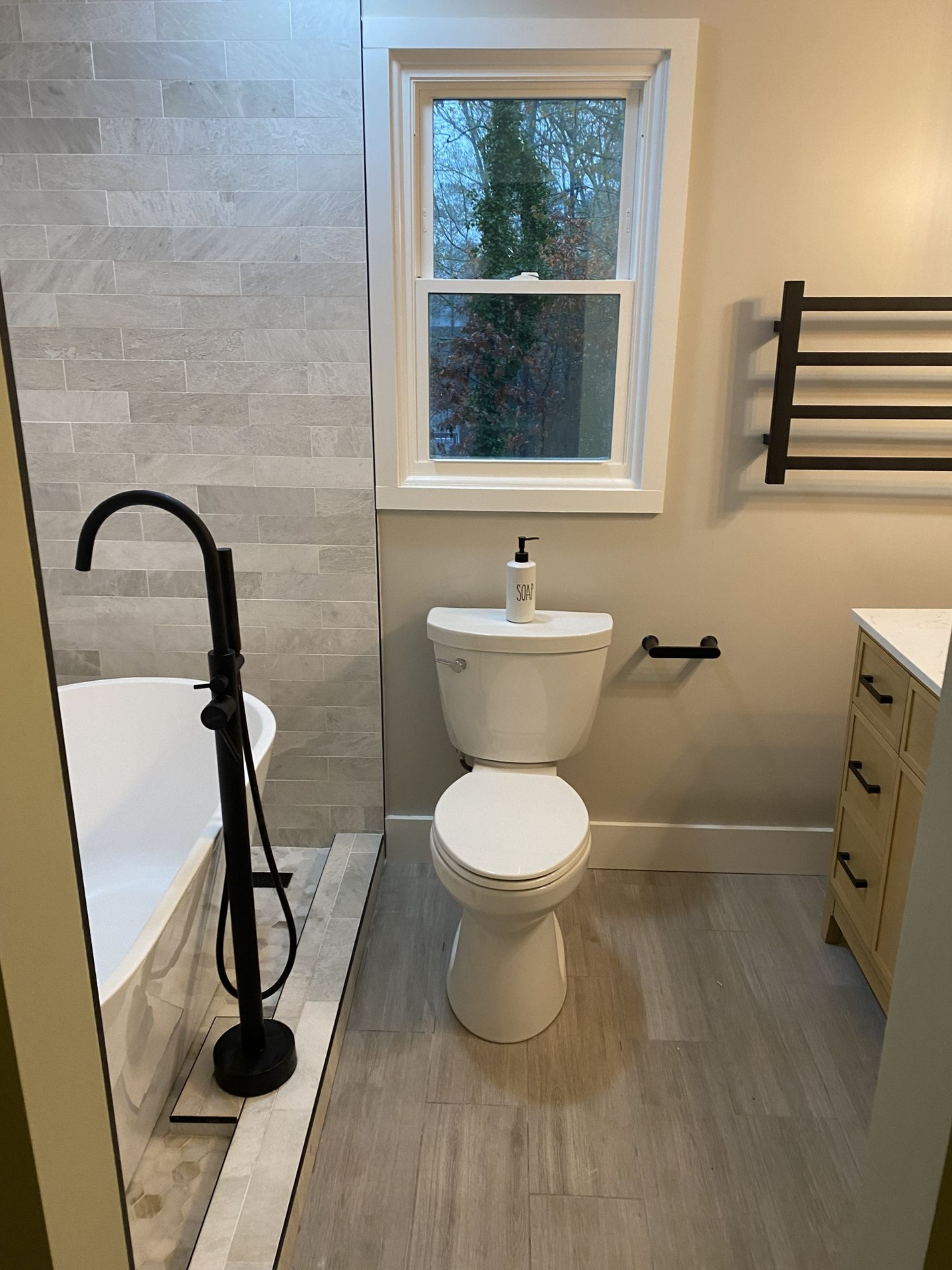 Bathroom with bathtub, toilet, and window; gray tile wall, black fixtures, light wood-look floor.
