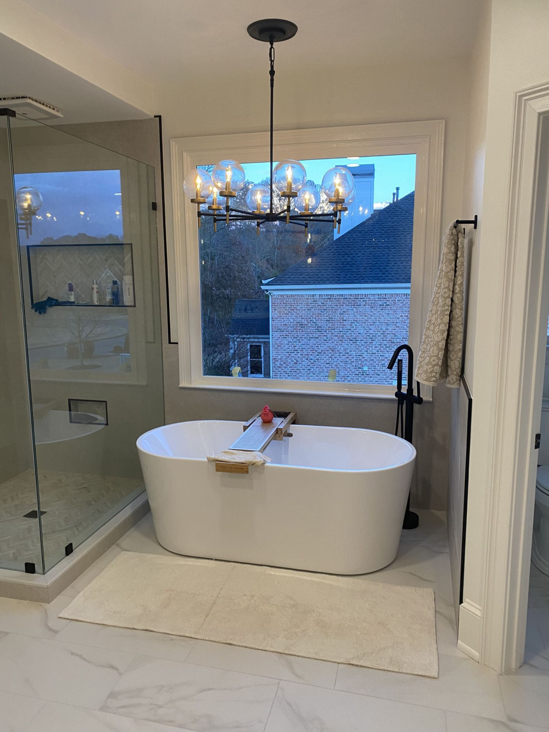 A modern bathroom with a white tub in front of a window. A shower and chandelier are also visible.