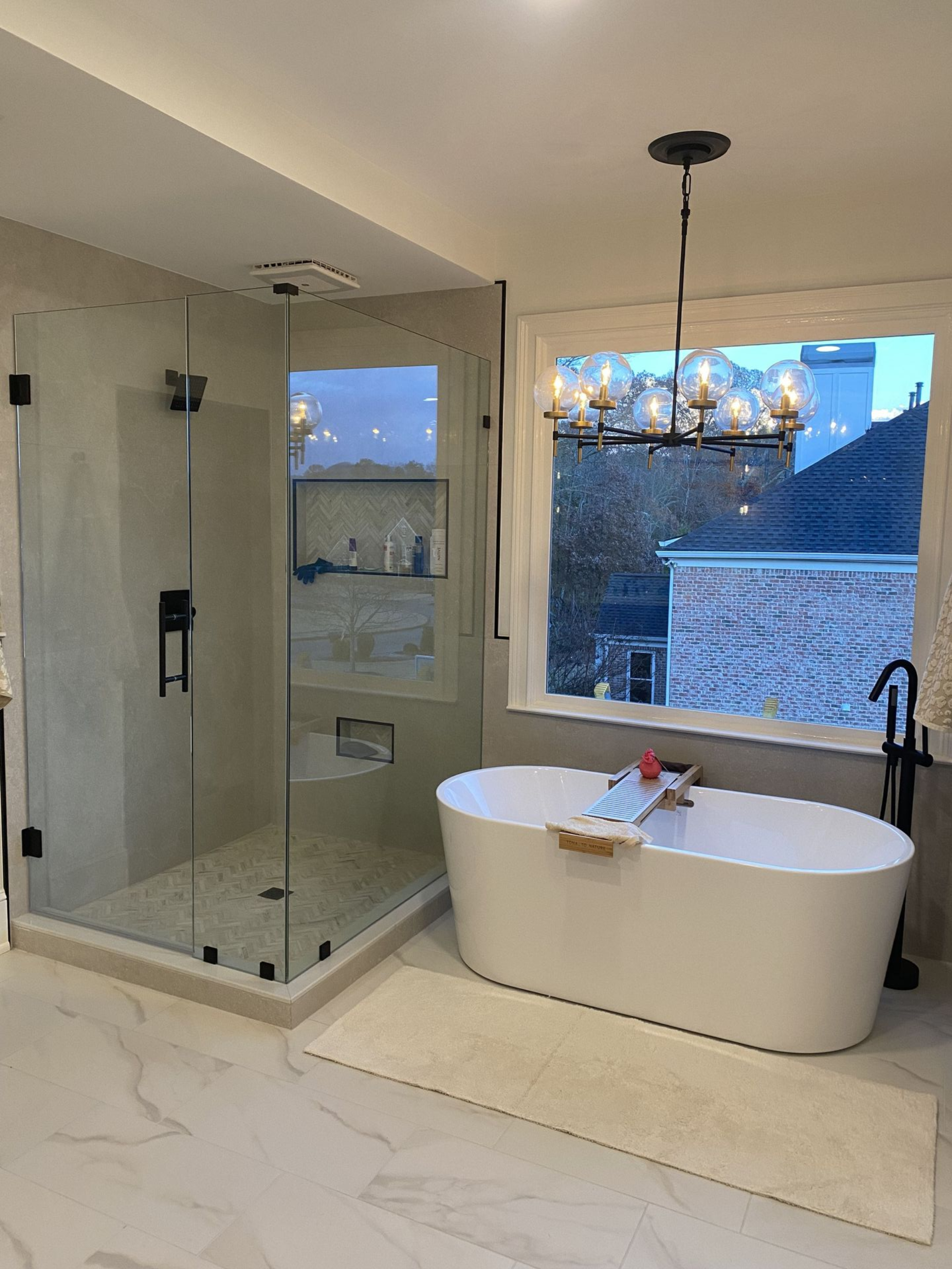 Modern bathroom with a glass shower, soaking tub, large window, and chandelier.