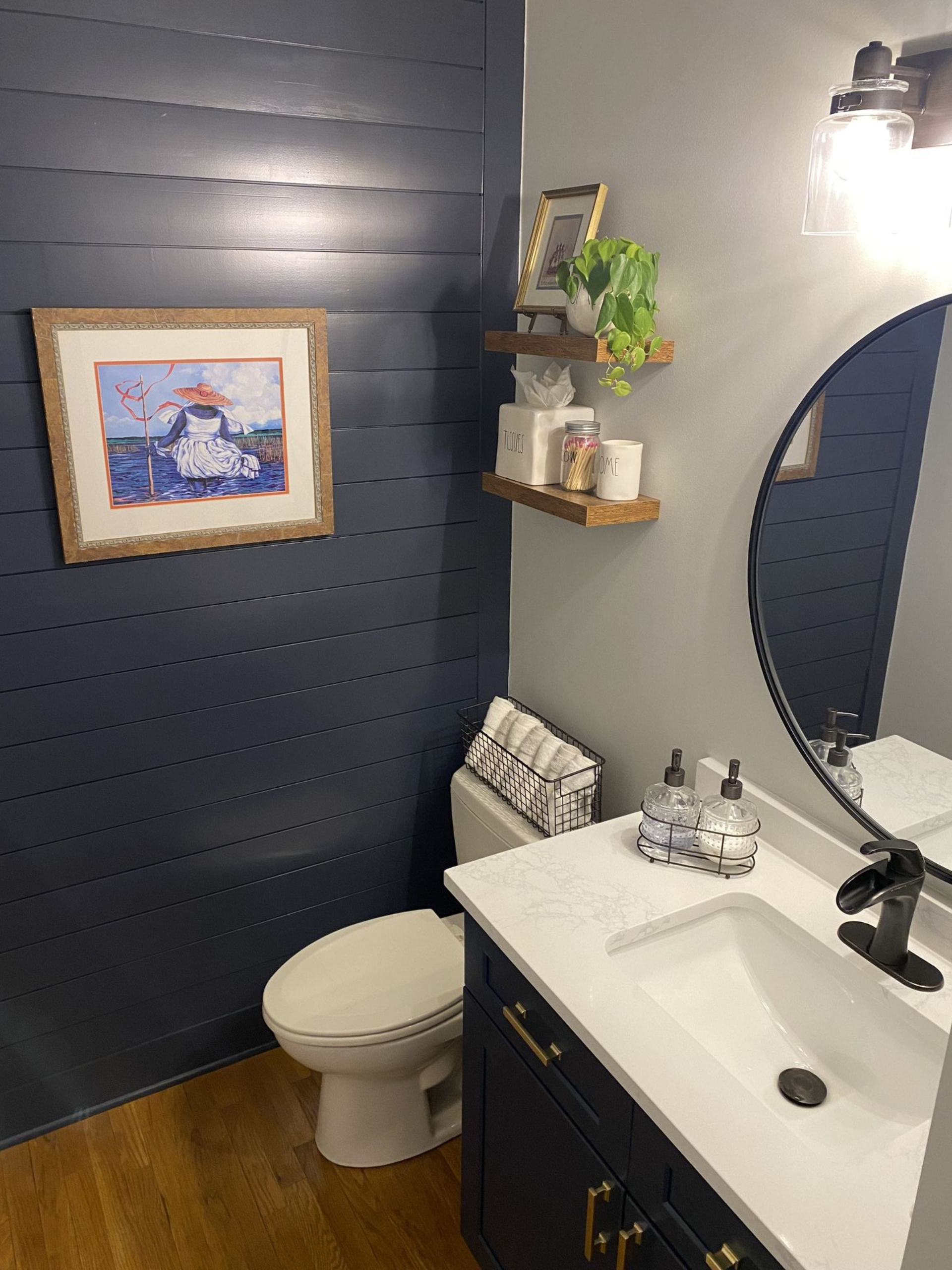 Navy blue and grey bathroom with toilet, sink, framed art, and wooden shelves.