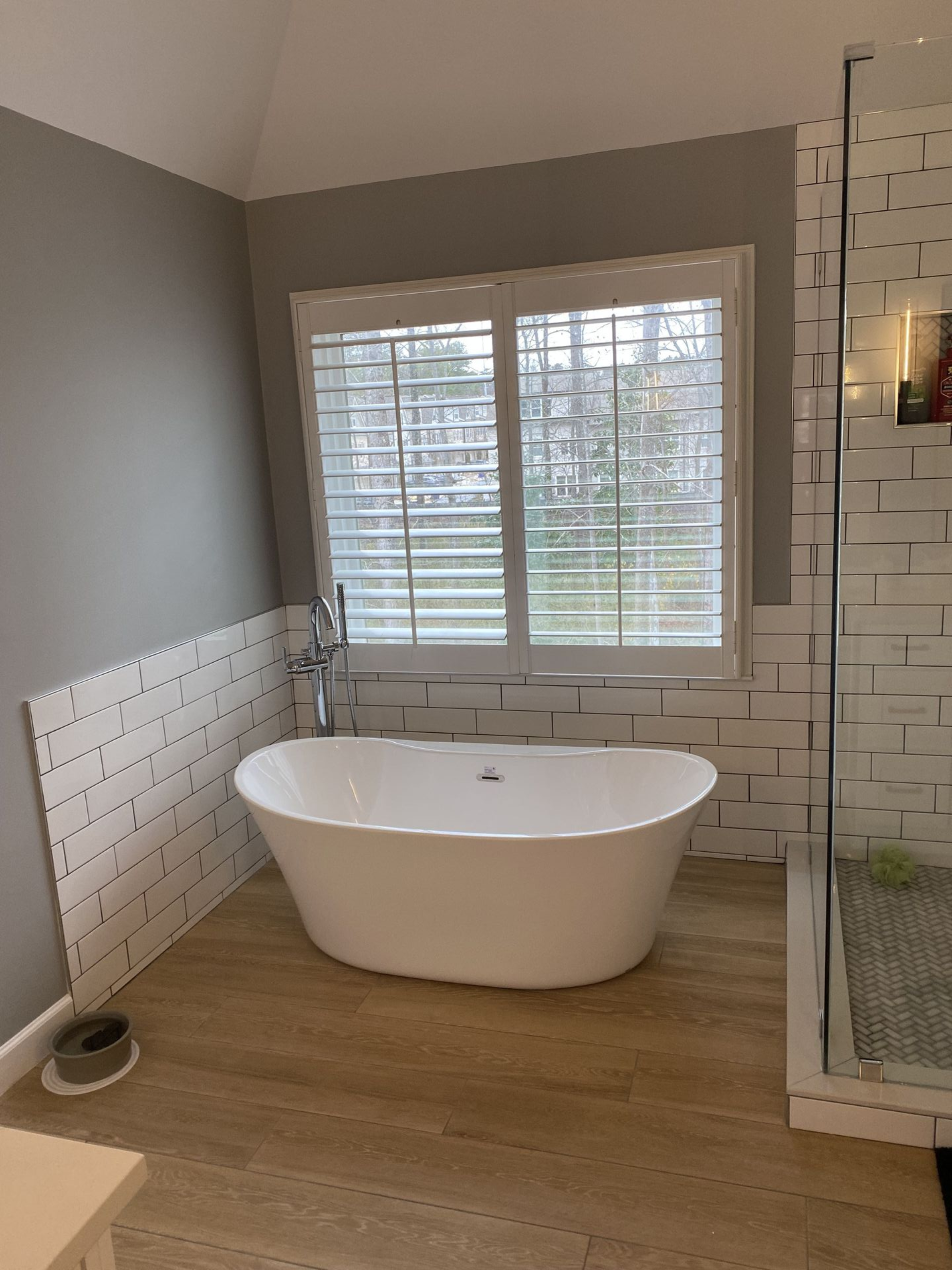 Bathroom with white soaking tub, light-colored flooring, window with shutters, and white tiled walls.