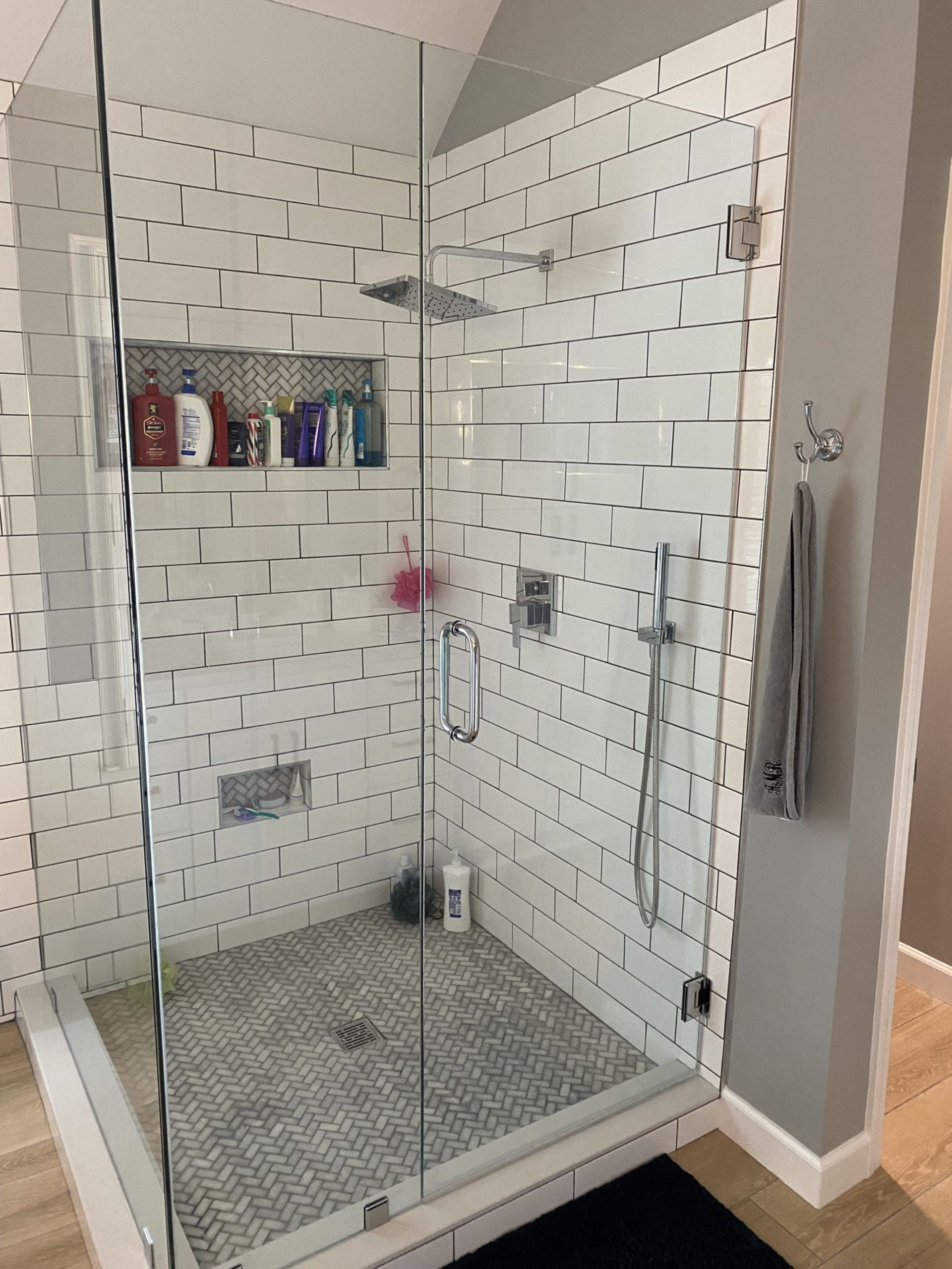 Glass-enclosed shower with white brick-like tiles, a built-in shelf with toiletries, and a gray patterned floor.