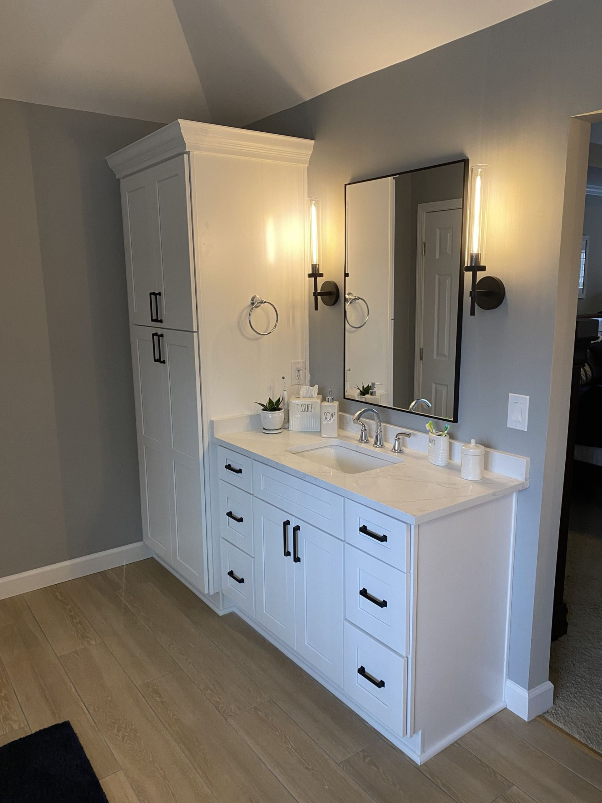 White bathroom vanity and tall storage cabinet with black hardware and light fixtures.