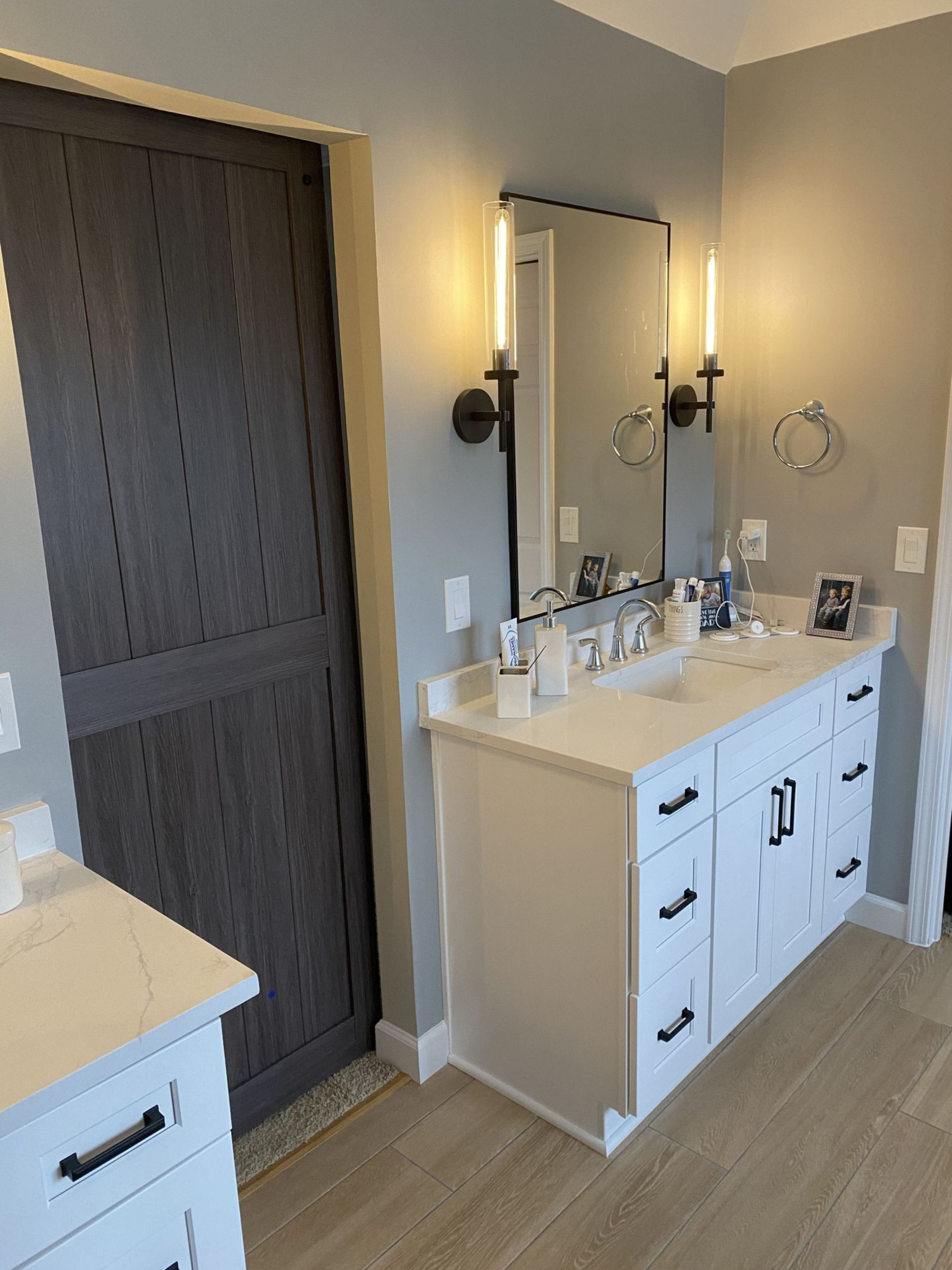 Bathroom with white vanity, large mirror, and dark gray door. Two black sconces flank the mirror on gray walls.