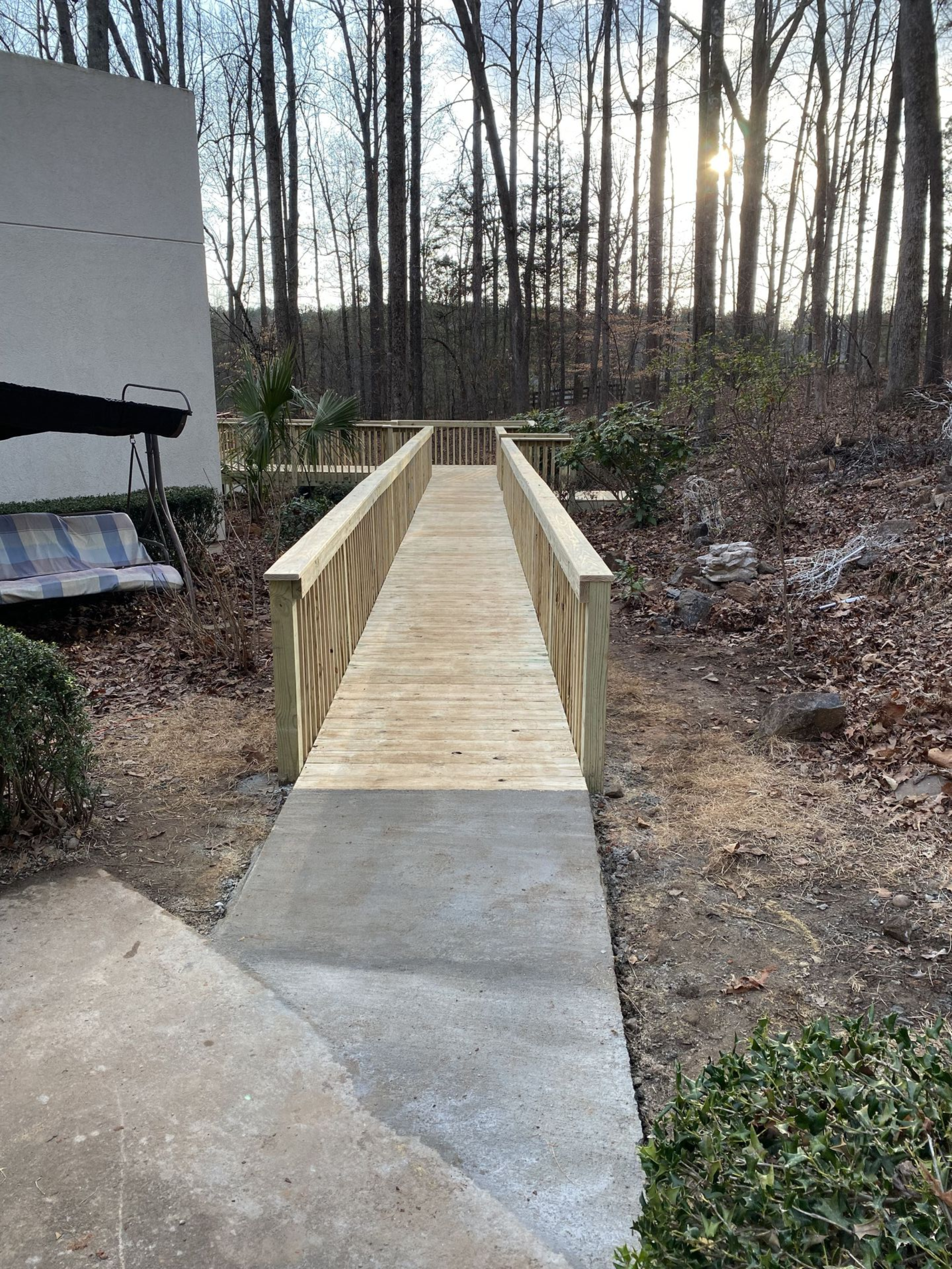 Wooden ramp over gravel and foliage, leading from a concrete path to trees; sunlight visible.