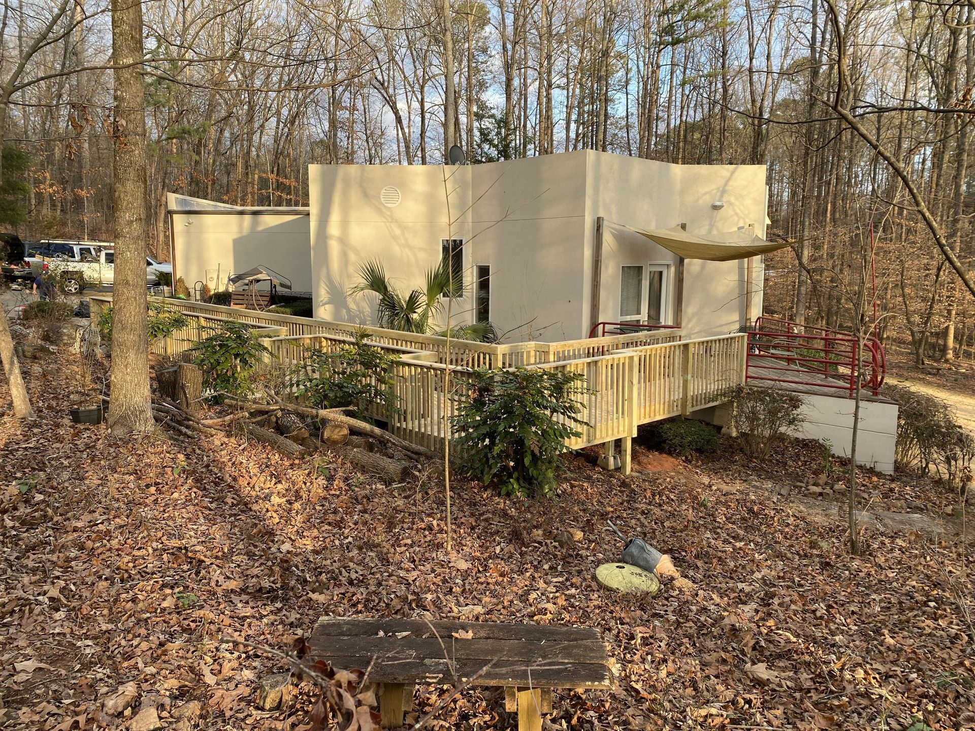 Beige house with a wooden deck in a wooded area. Dry leaves cover the ground.