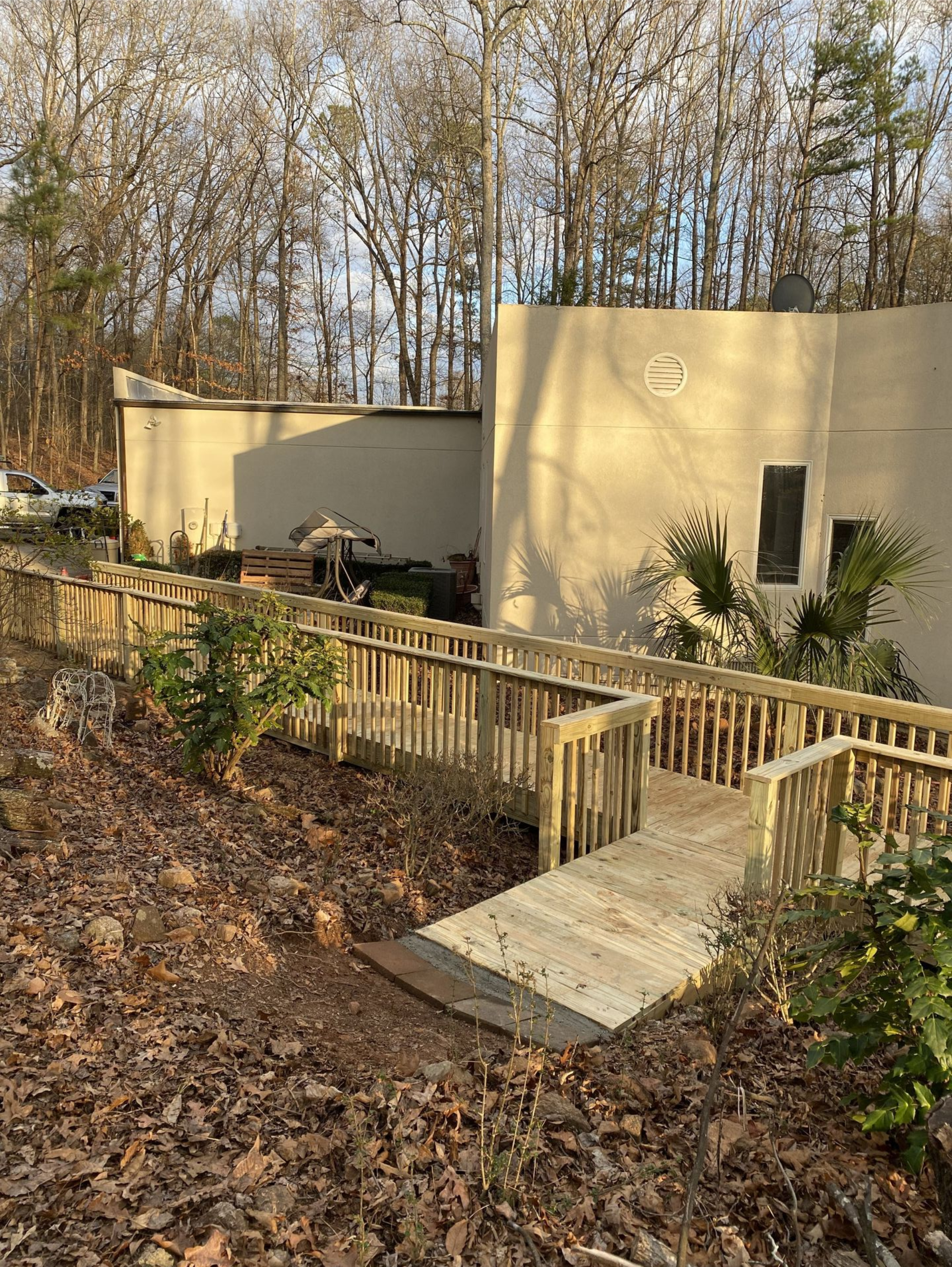 A wooden ramp leads to a tan building with a doorway. Bushes and trees surround the structure.