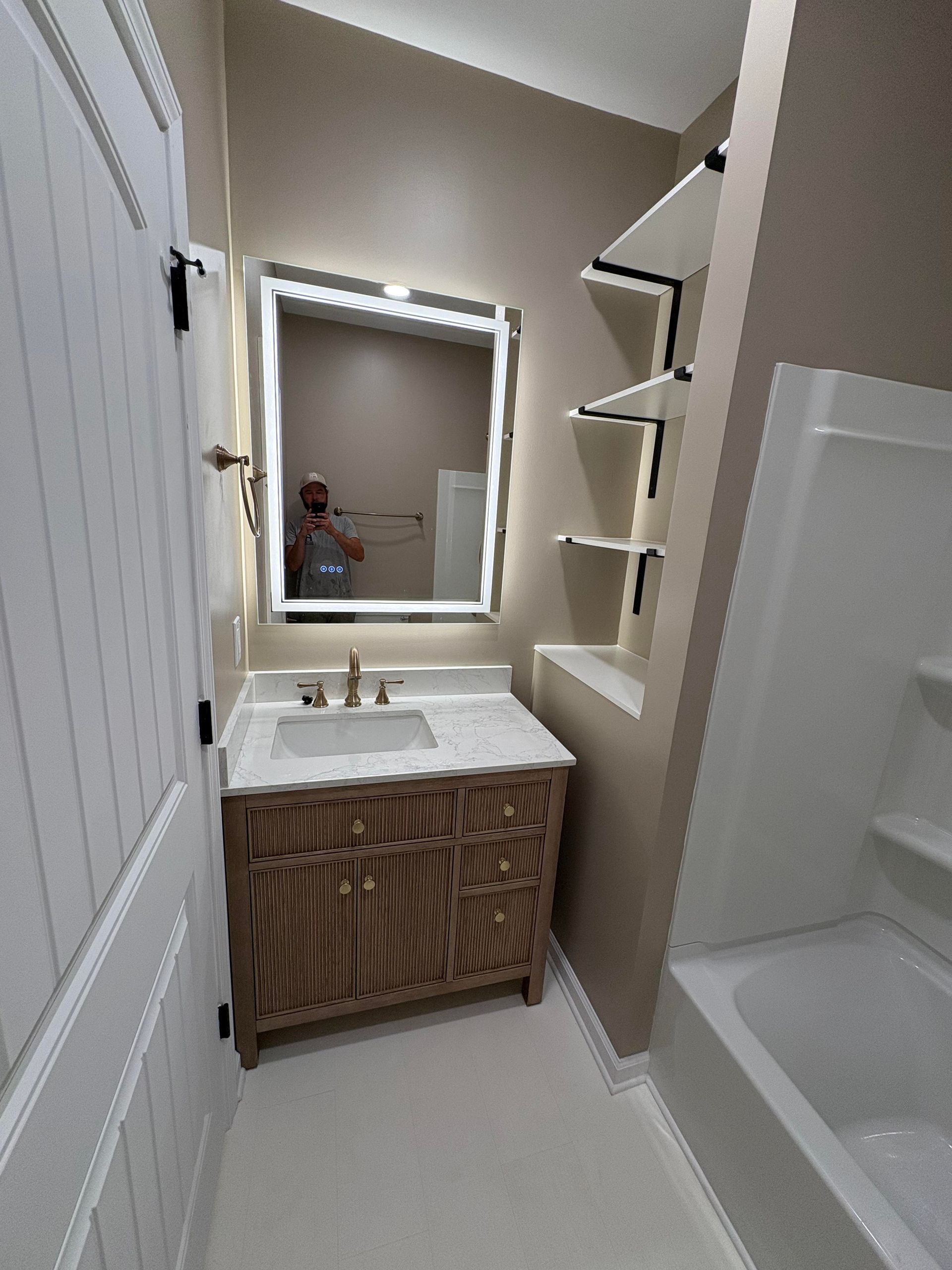Bathroom with vanity, LED mirror, shelving, and bathtub. Beige walls, wood vanity, white trim.