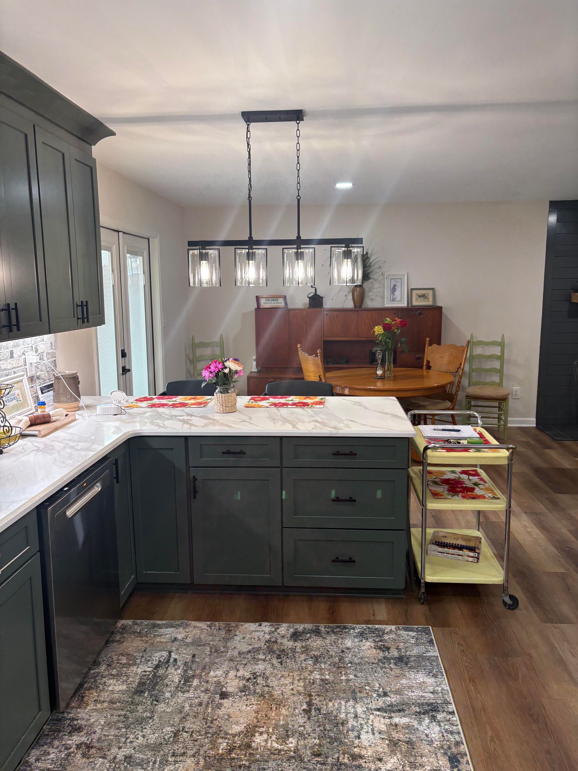 Green kitchen with island, white countertop, dark cabinets, rug, and pendant lights.