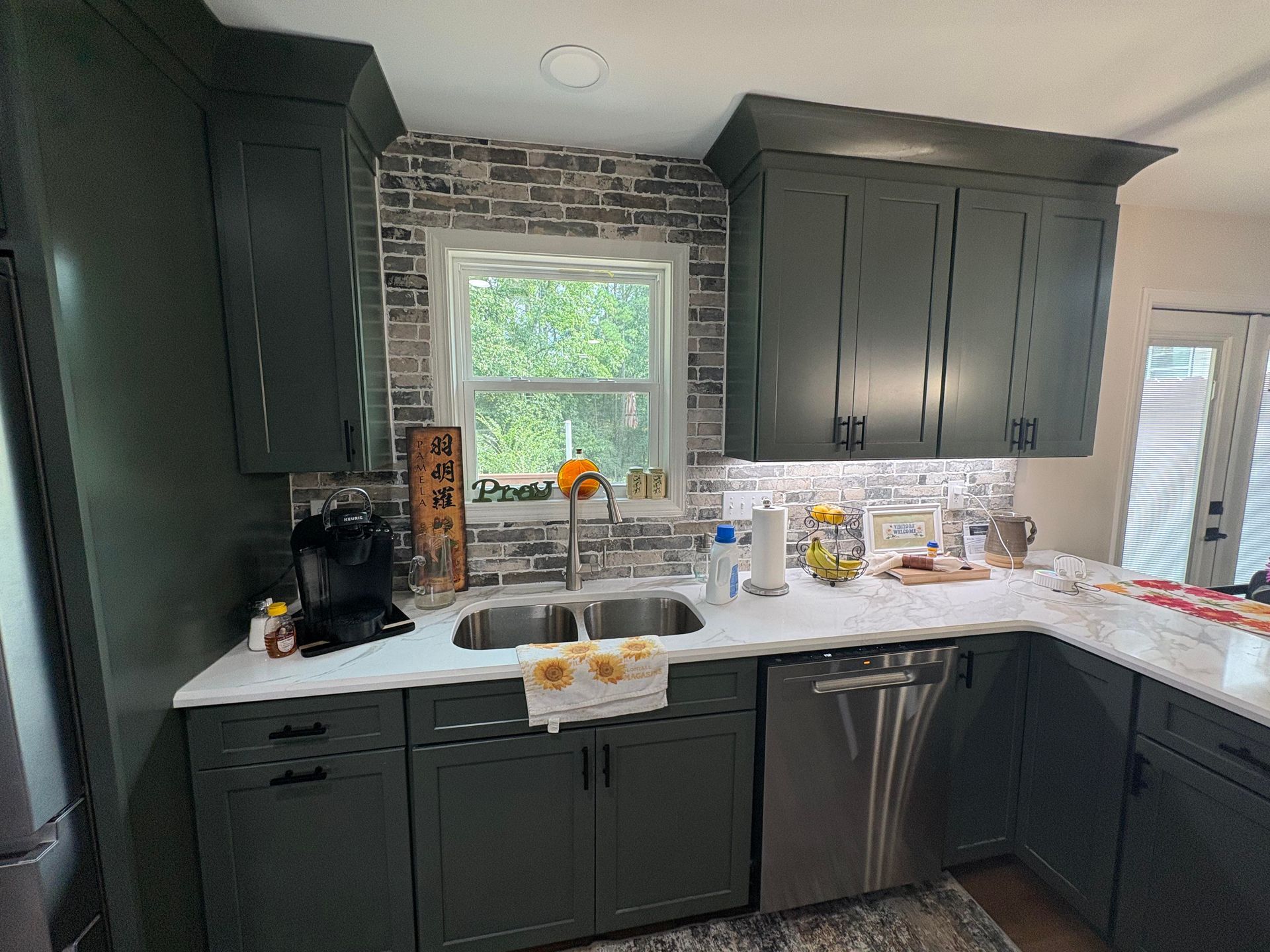 Dark green kitchen cabinets with white countertops, stainless steel appliances, and a brick backsplash.