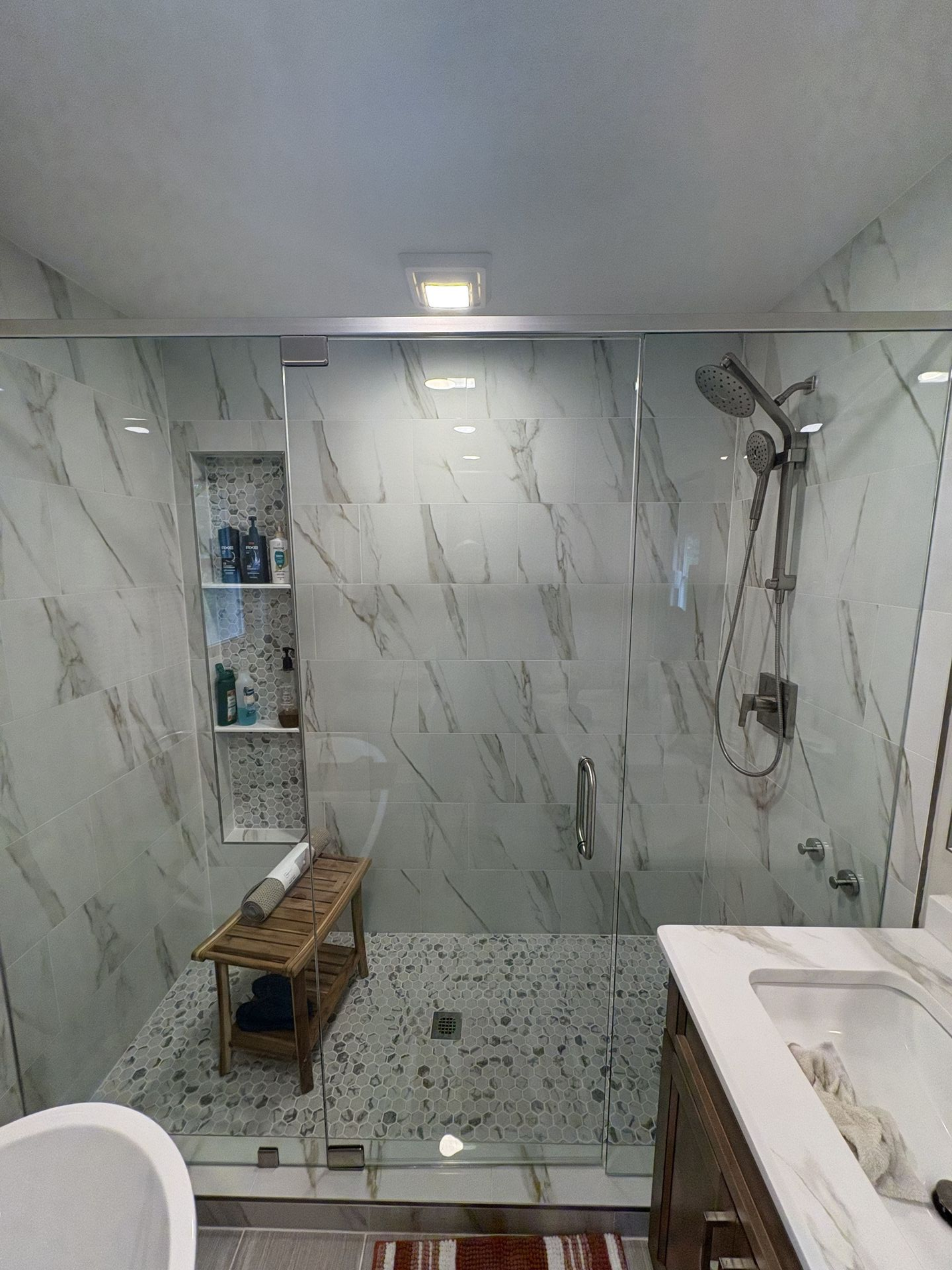 Bathroom with marble-look tile shower, glass door, built-in shelving, wooden bench, and vanity with sink.