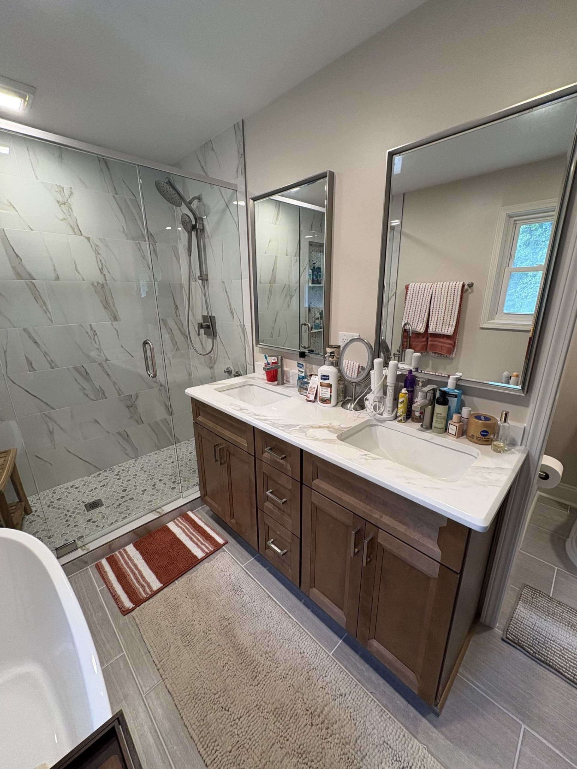 Bathroom with a double vanity, marble shower, and large mirrors. Brown cabinets, gray and white tile, and a bathtub visible.