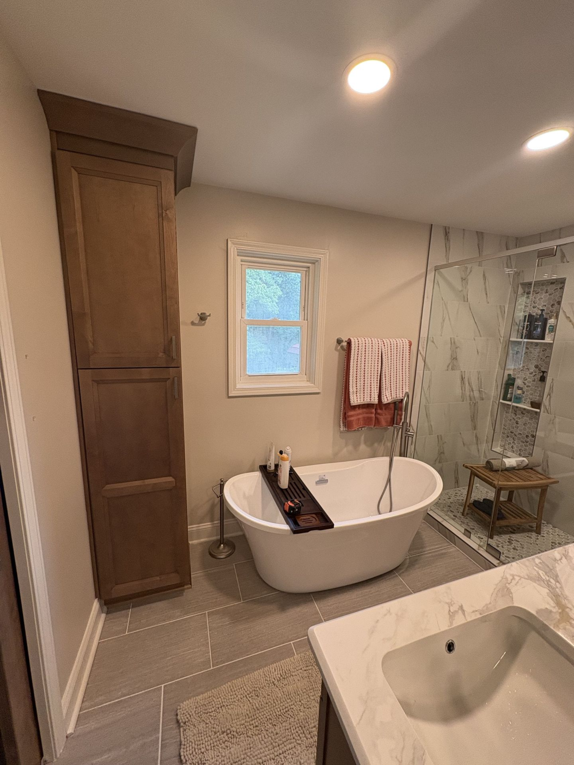 Bathroom with a freestanding tub, cabinet, window, and marble-look shower. Beige and gray color scheme.