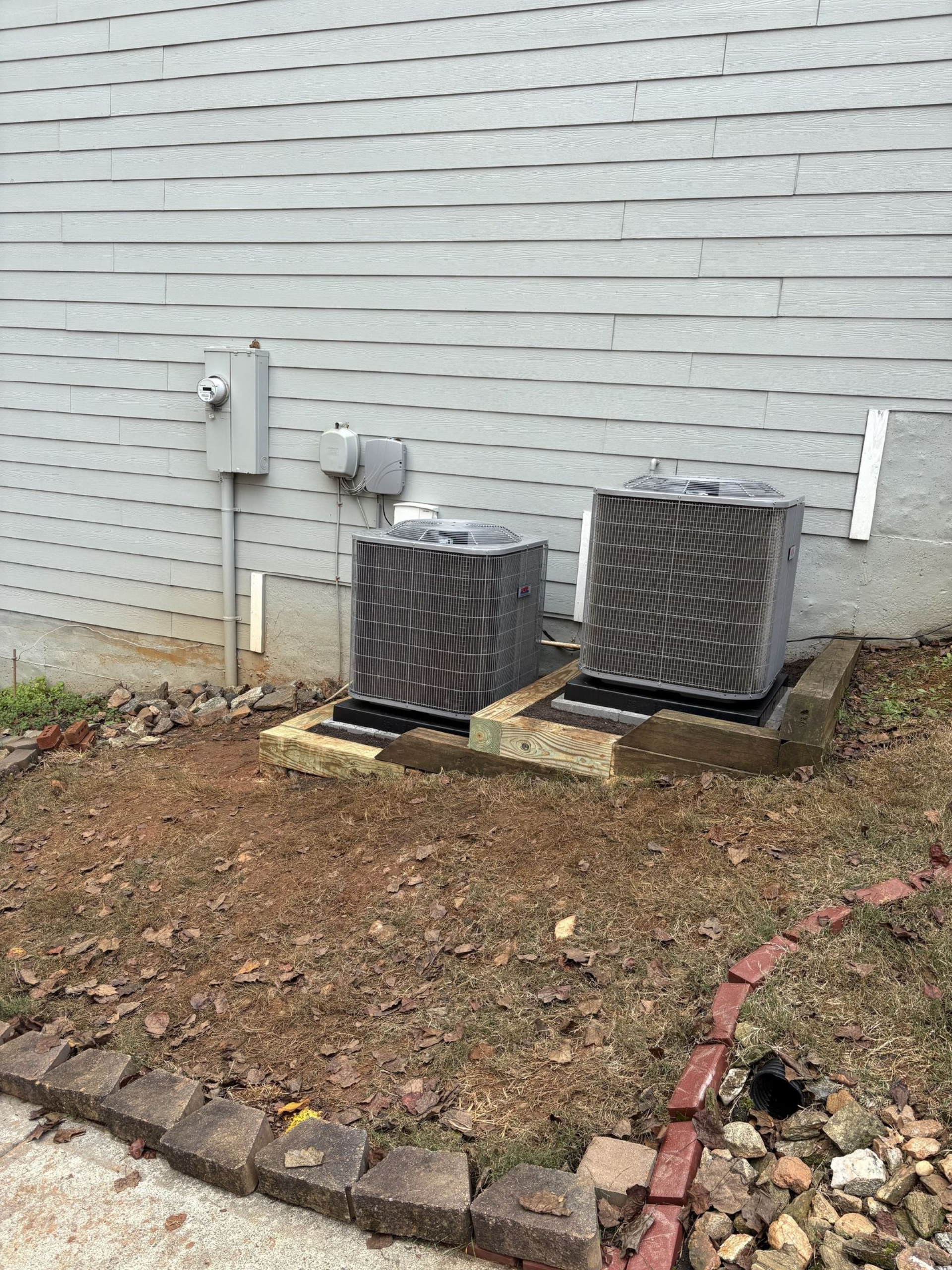 Two outdoor air conditioning units on concrete pads near a building with gray siding and electrical boxes.