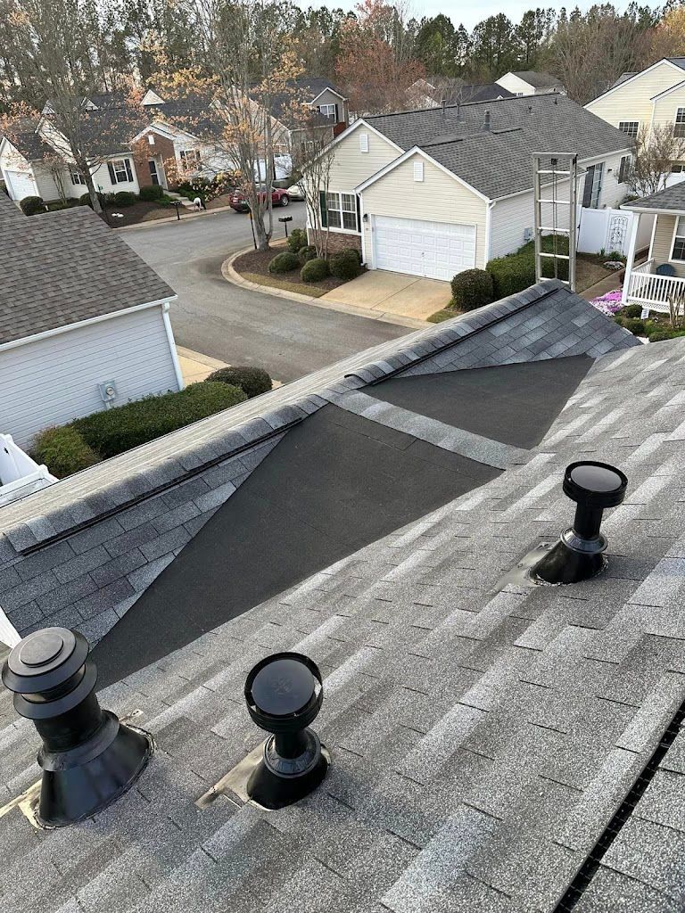 Rooftop view of a house with gray shingles.  Dark vent pipes and a patch of black material.  Suburban setting.