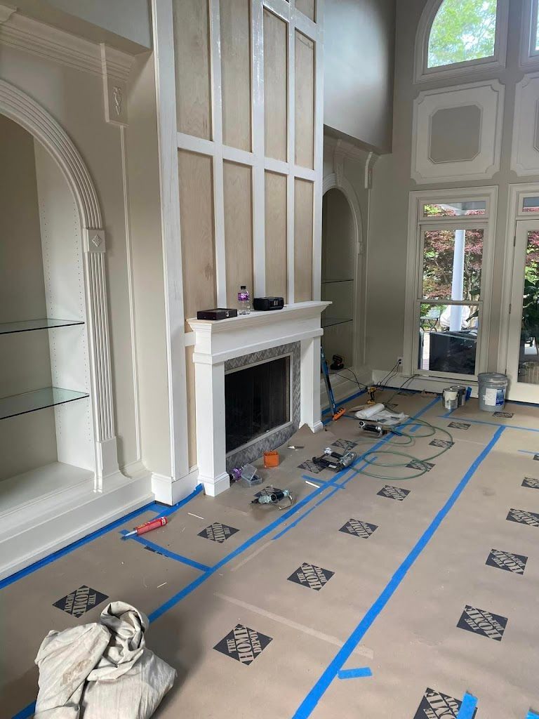 Interior view of a living room under renovation. Fireplace, built-in shelves, and windows are visible. Blue tape on floor.