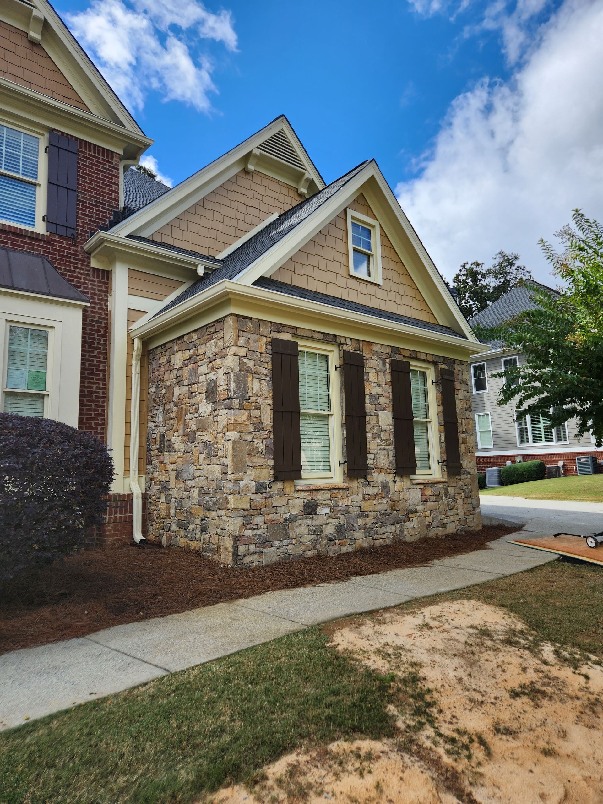 Stone-clad house with brown shutters, tan siding, and a small gable window. Sunny day.