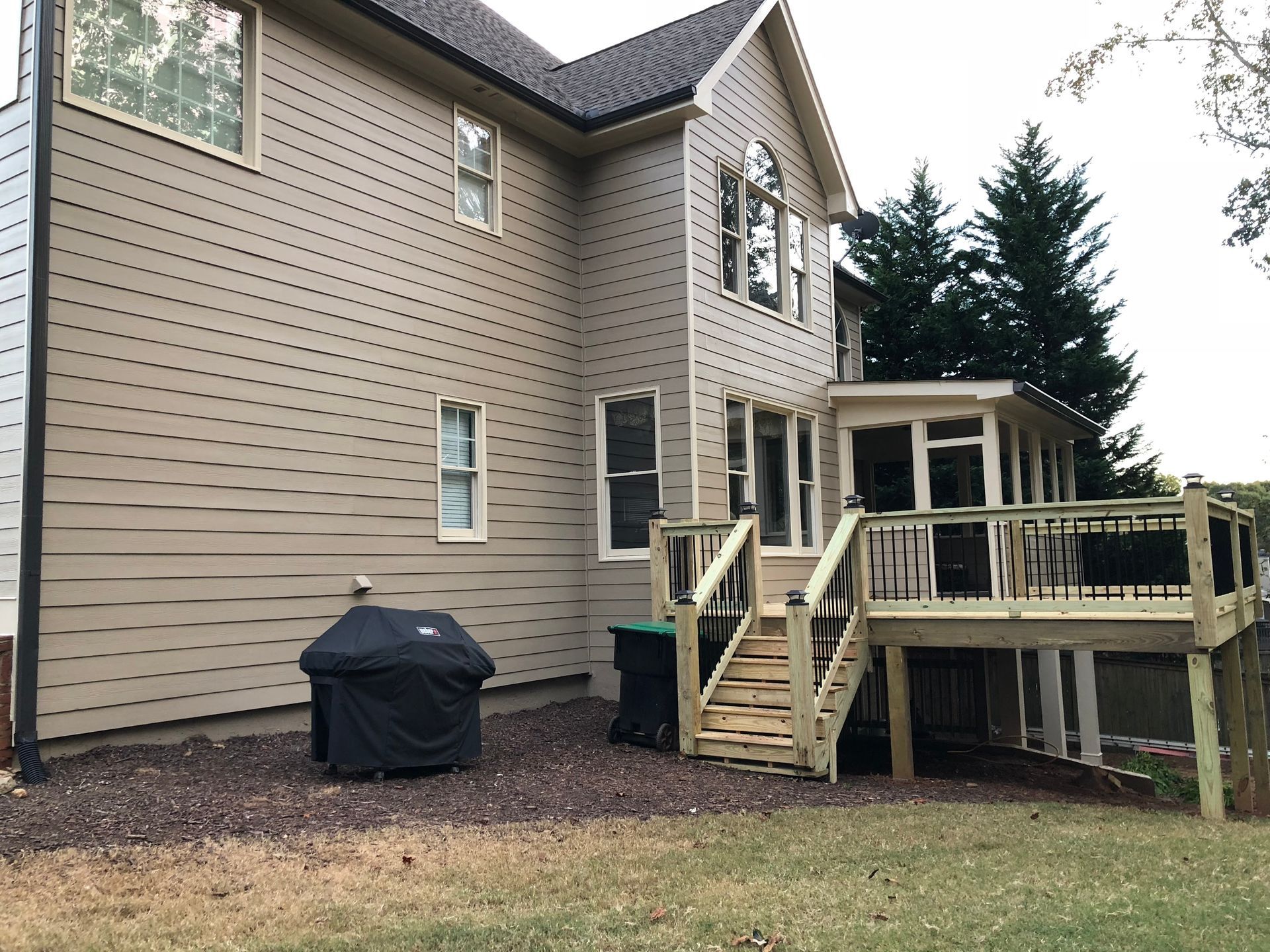 Backyard of a two-story beige house with a wooden deck and a black grill on a gravel bed.