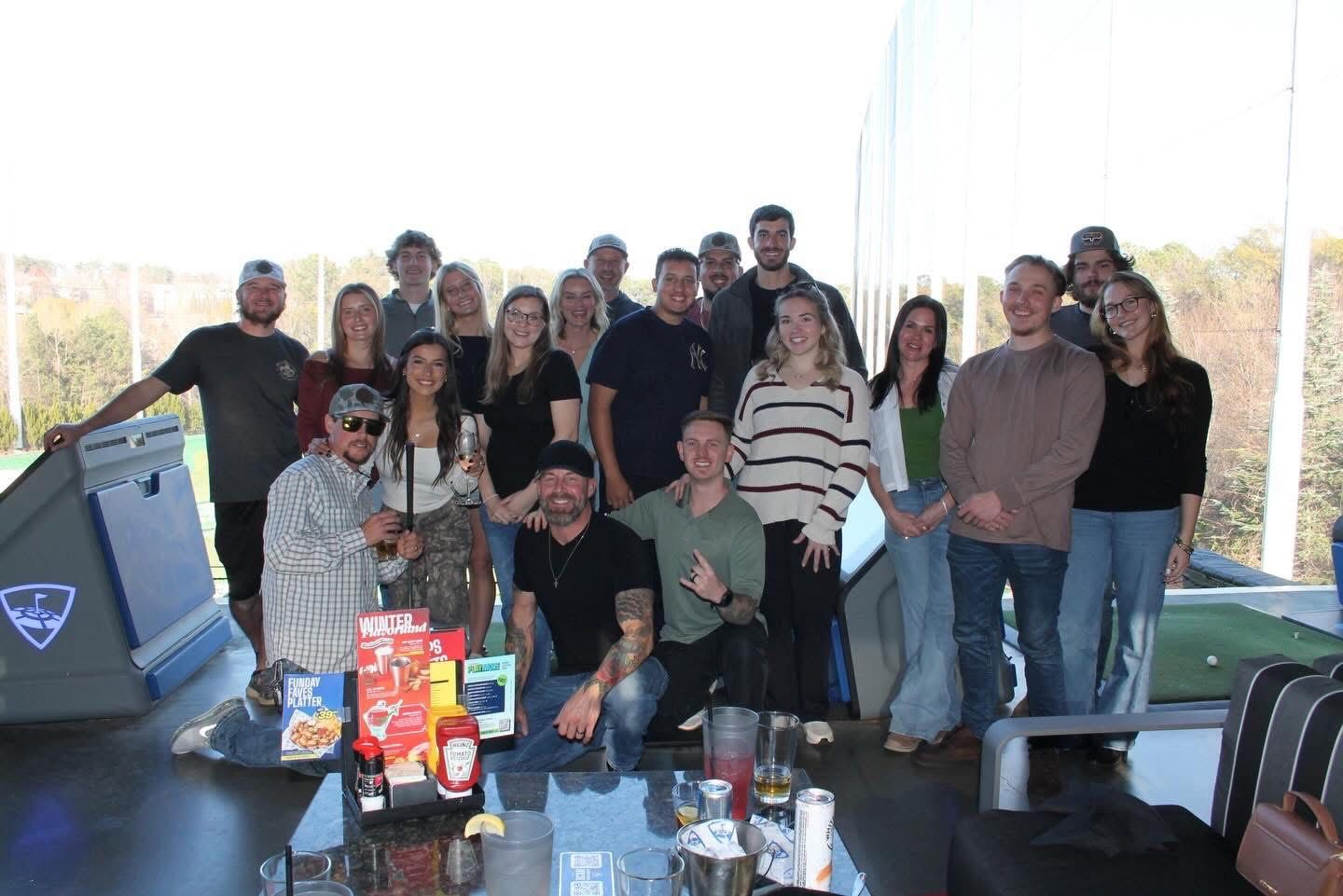 Group of people at a Topgolf, posing with drinks. Sunny day.