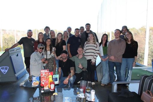 Group of people at a Topgolf, posing near a table with drinks and food. Blue skies, sunny day.