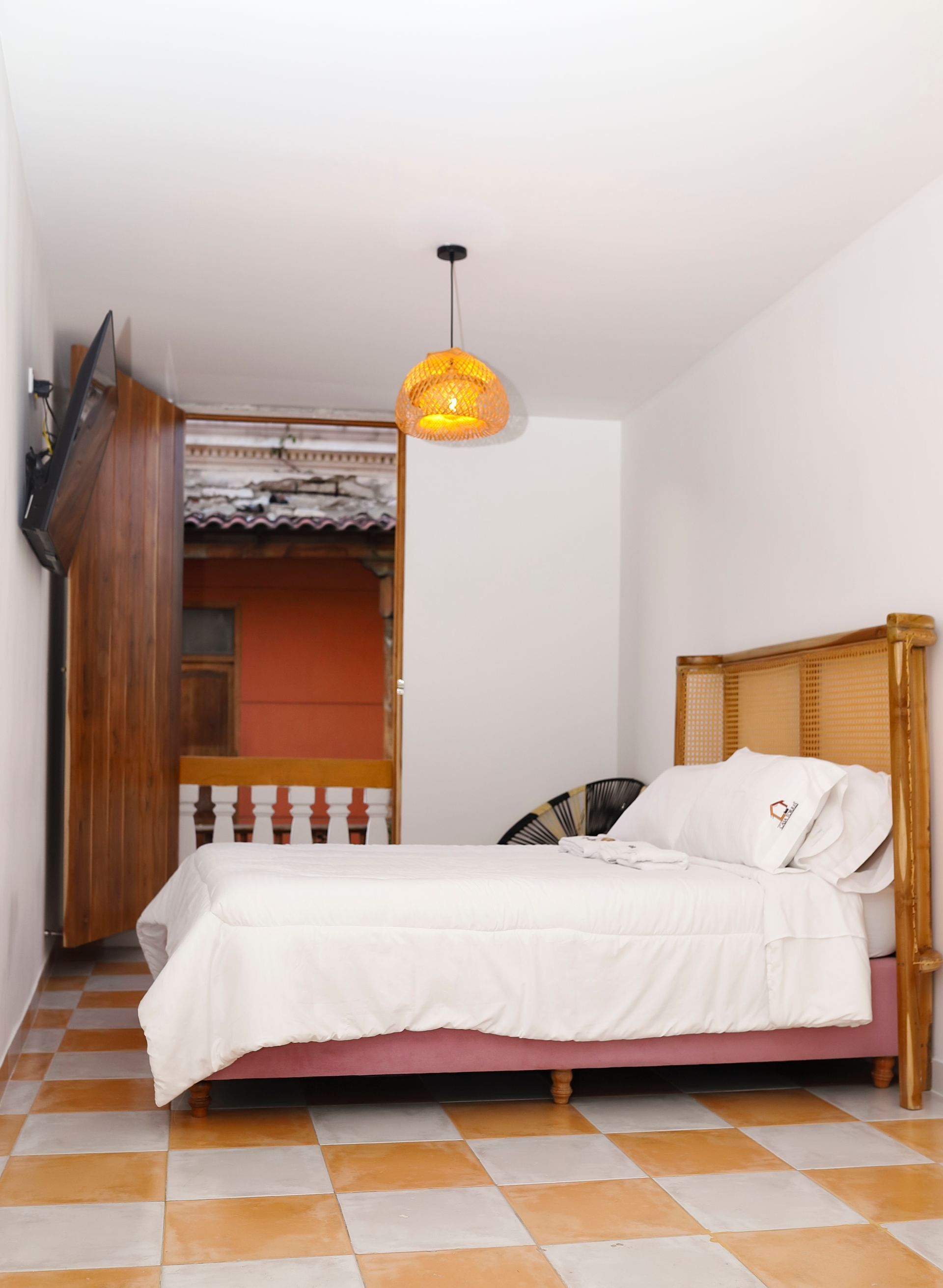 Dormitorio con cama, televisión, puerta de madera abierta a un balcón exterior y lámpara colgante.