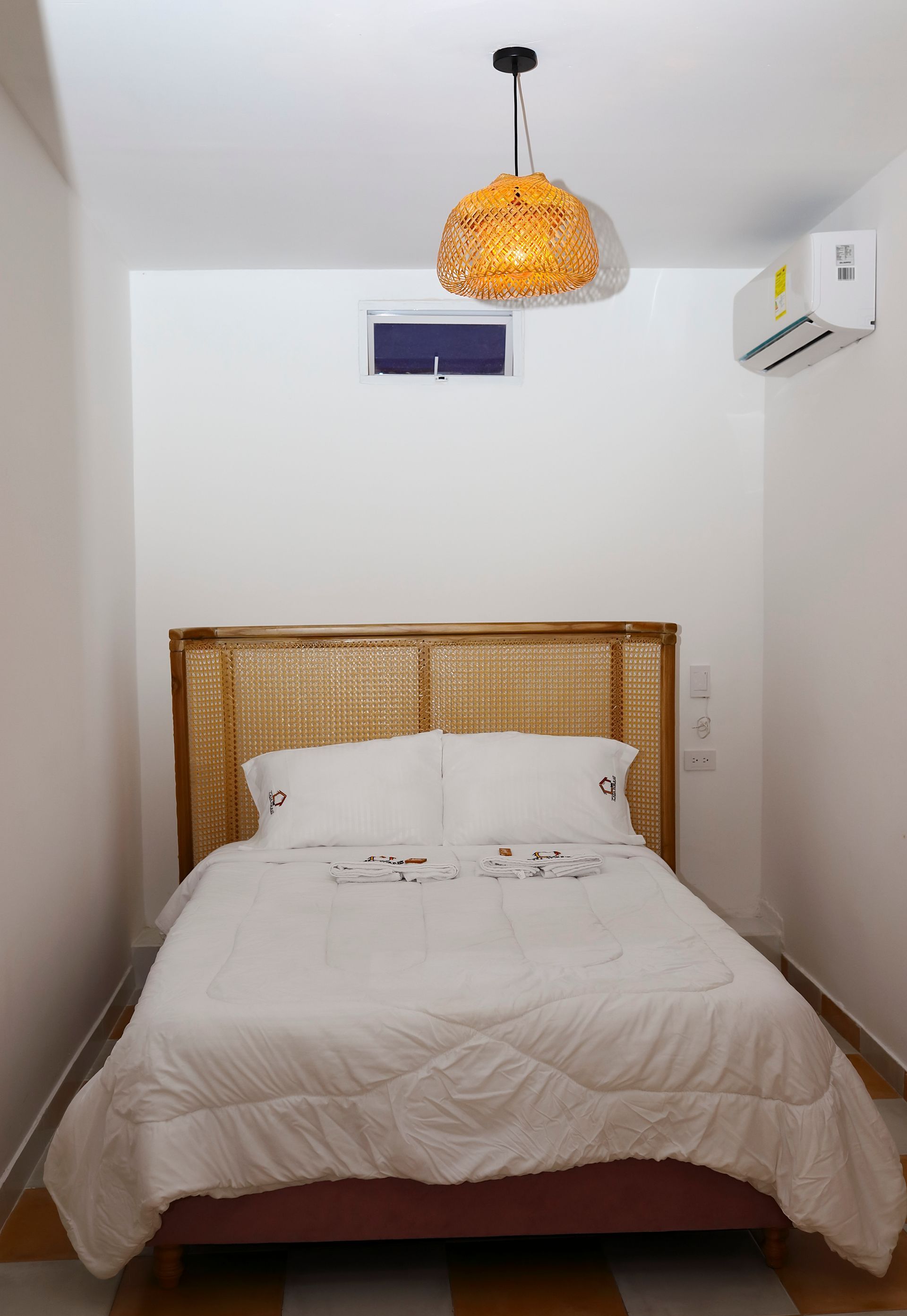 Dormitorio acogedor con cama, cabecero de mimbre, lámpara colgante y aire acondicionado. Paredes blancas, luminoso.