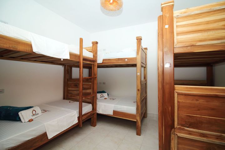 Literas en una habitación de paredes blancas con decoración sencilla. Estructuras de madera, ropa de cama blanca y una lámpara de techo decorativa.
